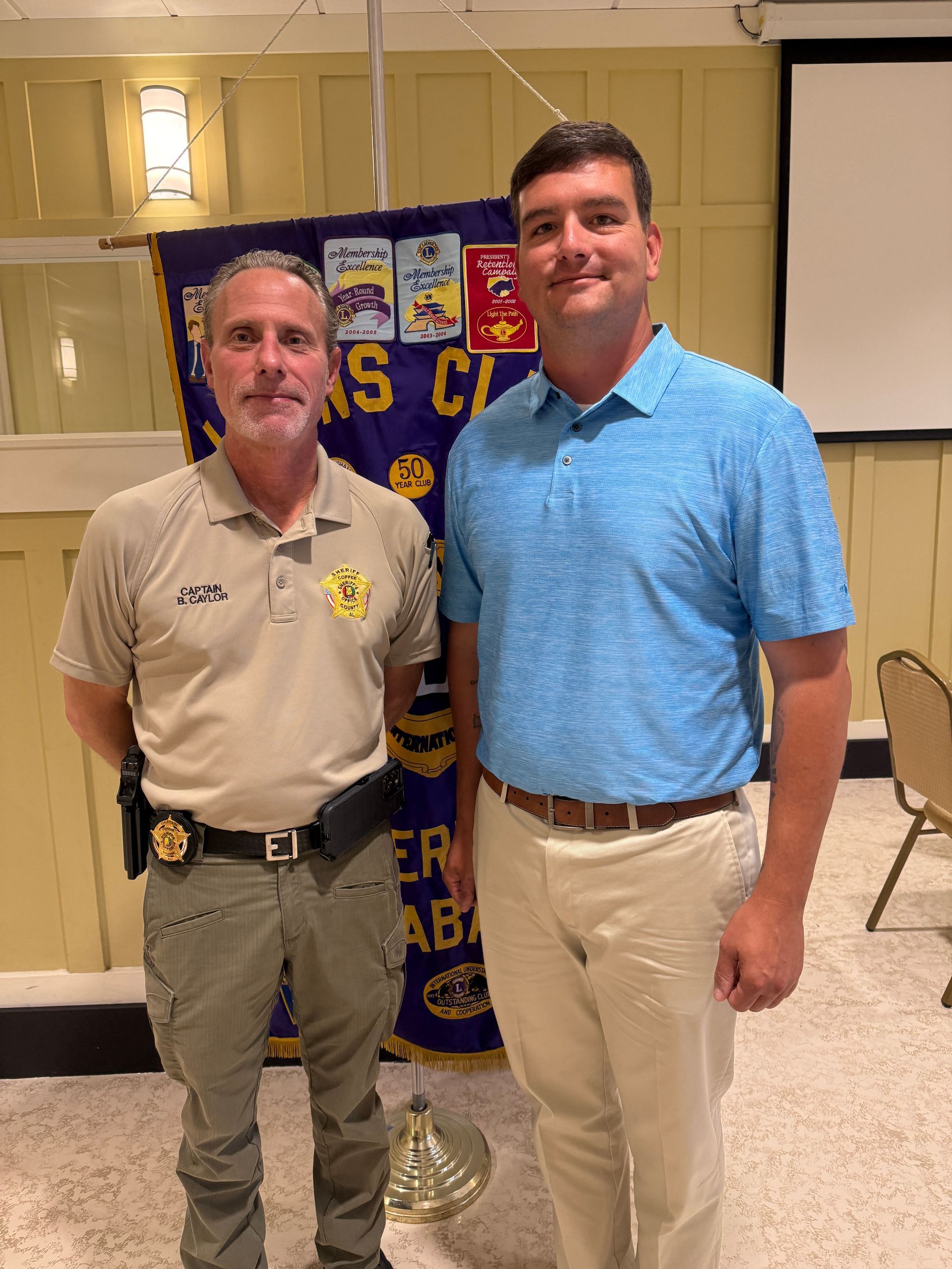 Two men standing indoors, one in a tan uniform and one in a blue polo, posing in front of a banner.