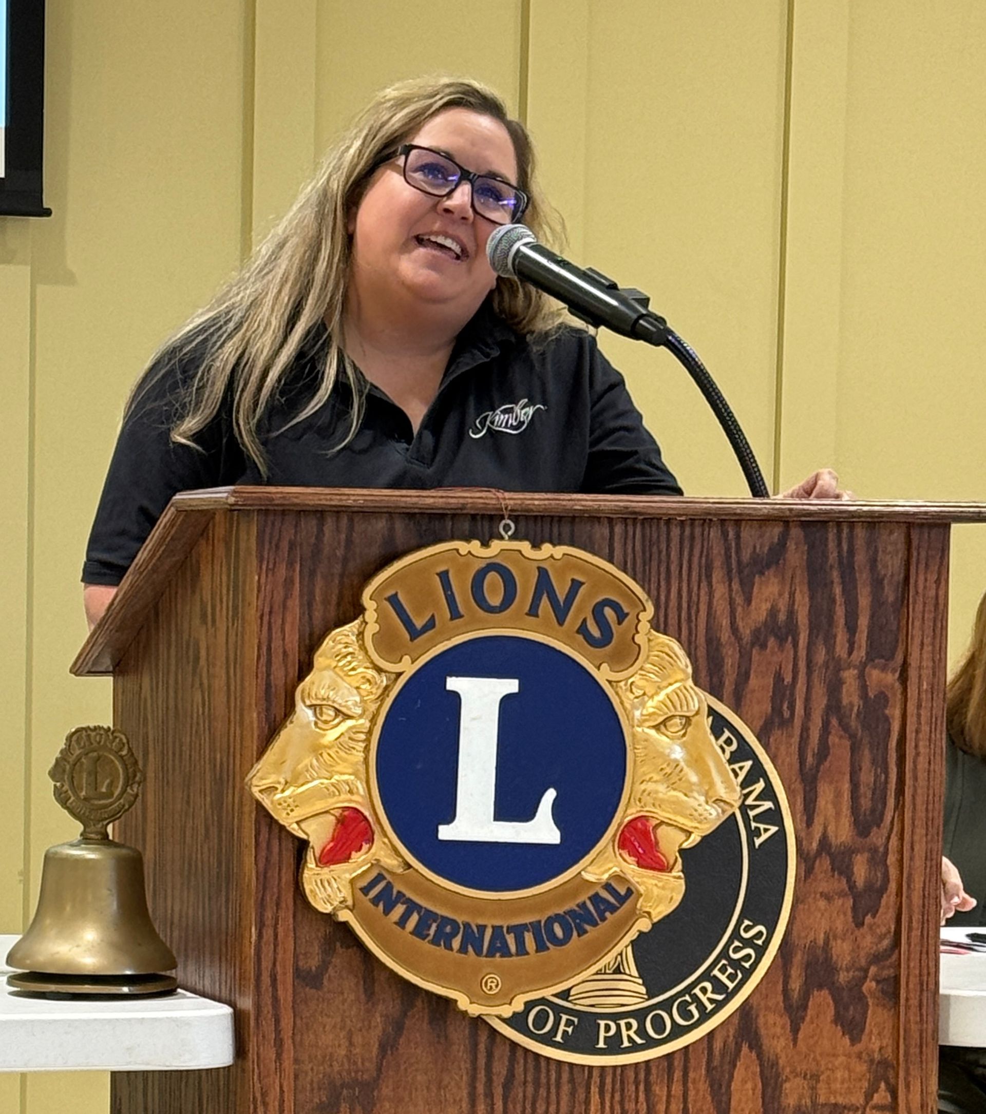 Woman speaking at a podium with a Lions Club emblem, smiling at a microphone in a conference room.