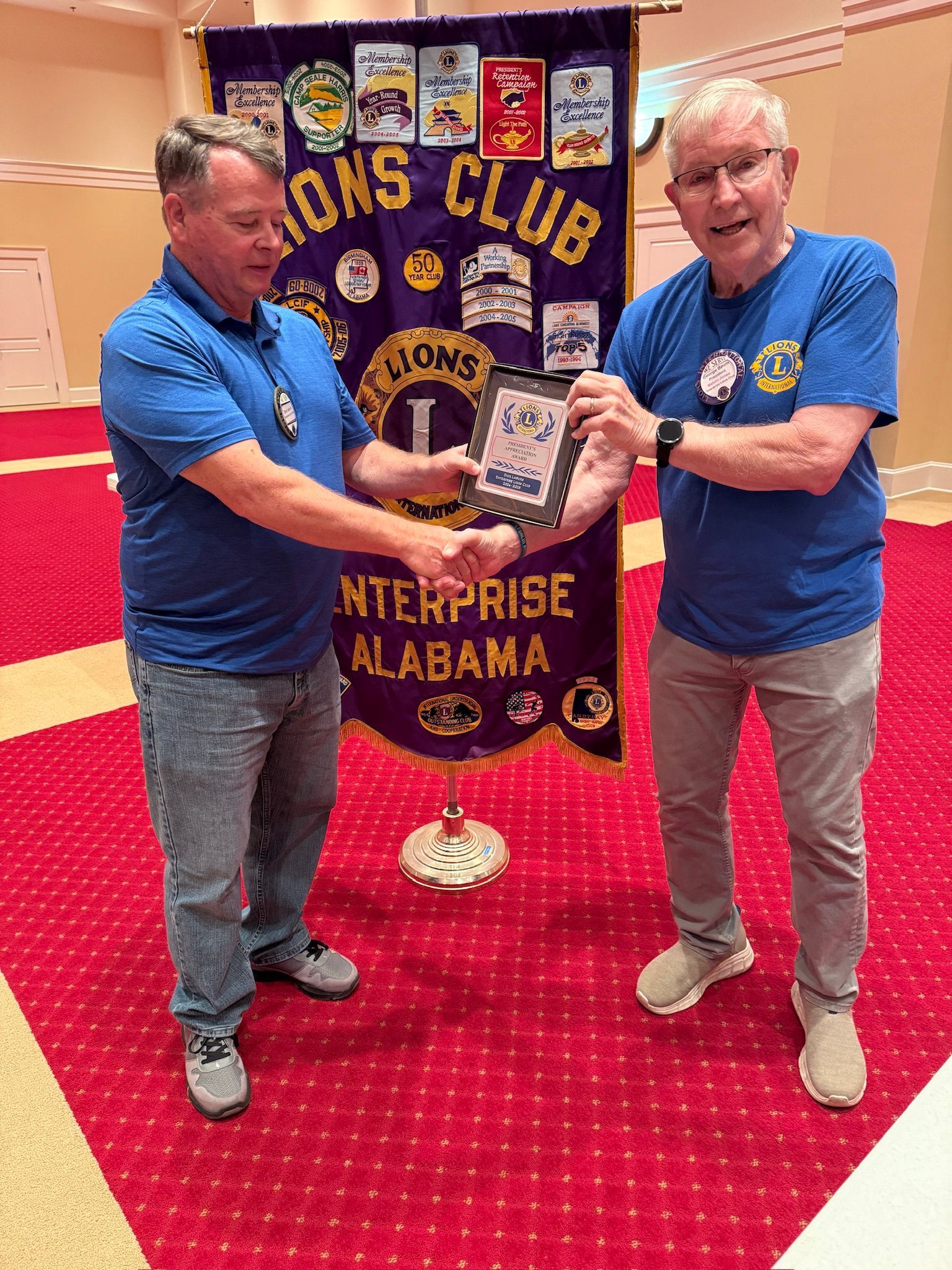 Two men in blue shirts shaking hands, receiving an award in front of a Lions Club banner in Enterprise, Alabama.