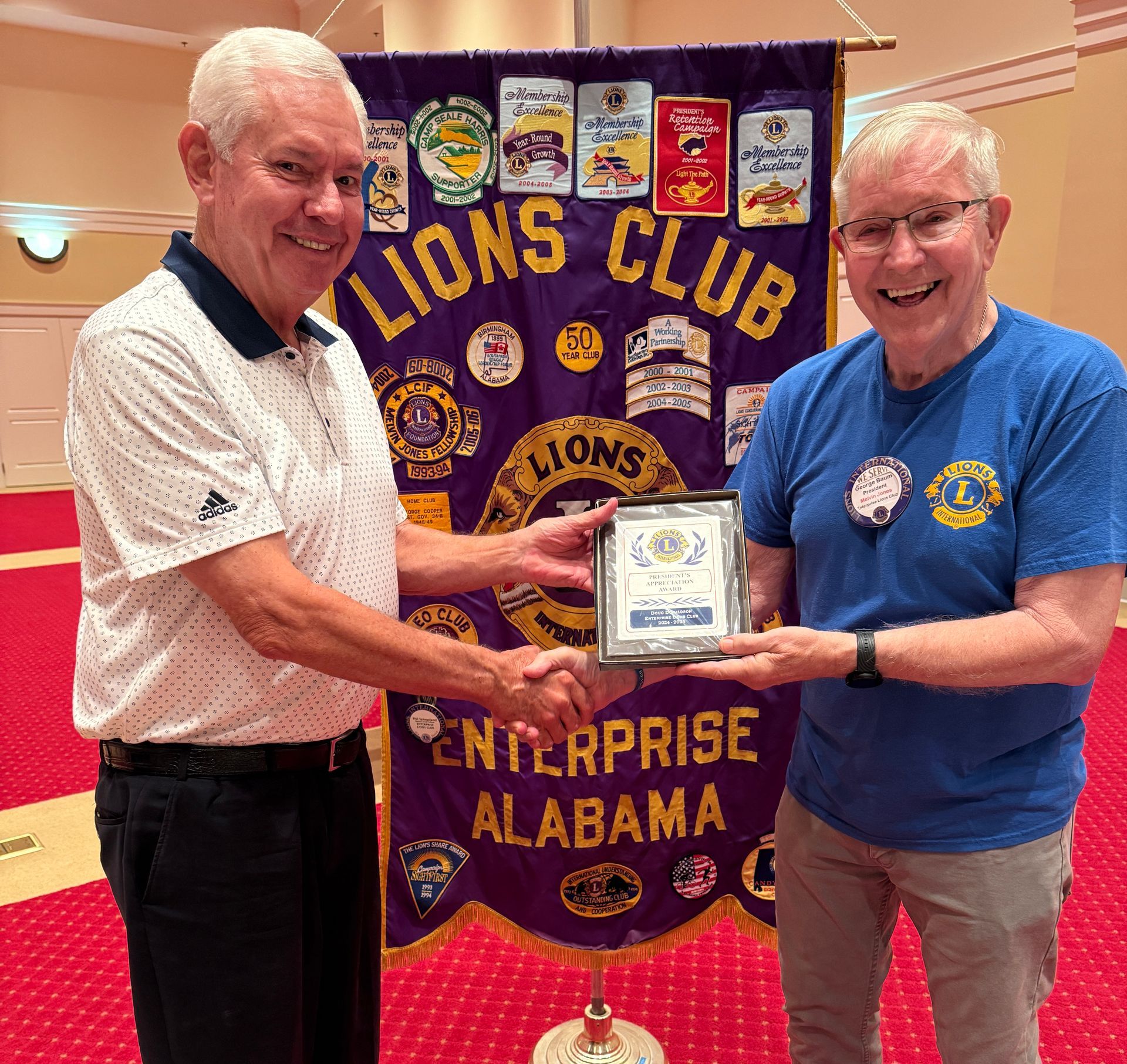 Two men shaking hands, holding a plaque in front of a Lions Club banner in Enterprise, Alabama.