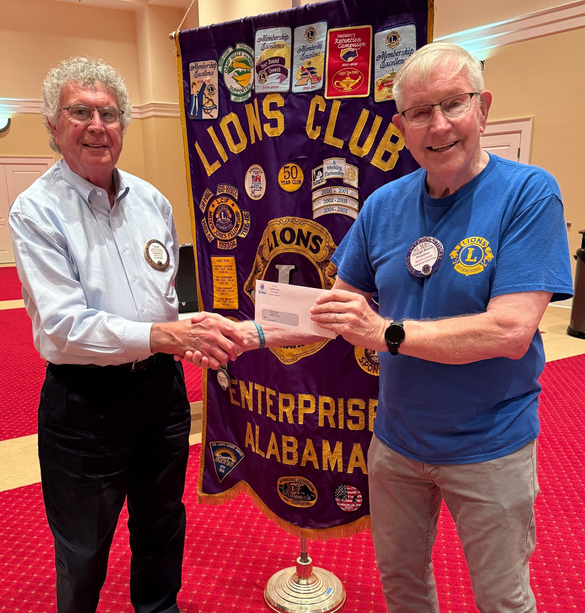 Two men shaking hands in front of a Lions Club banner. One hands a check.