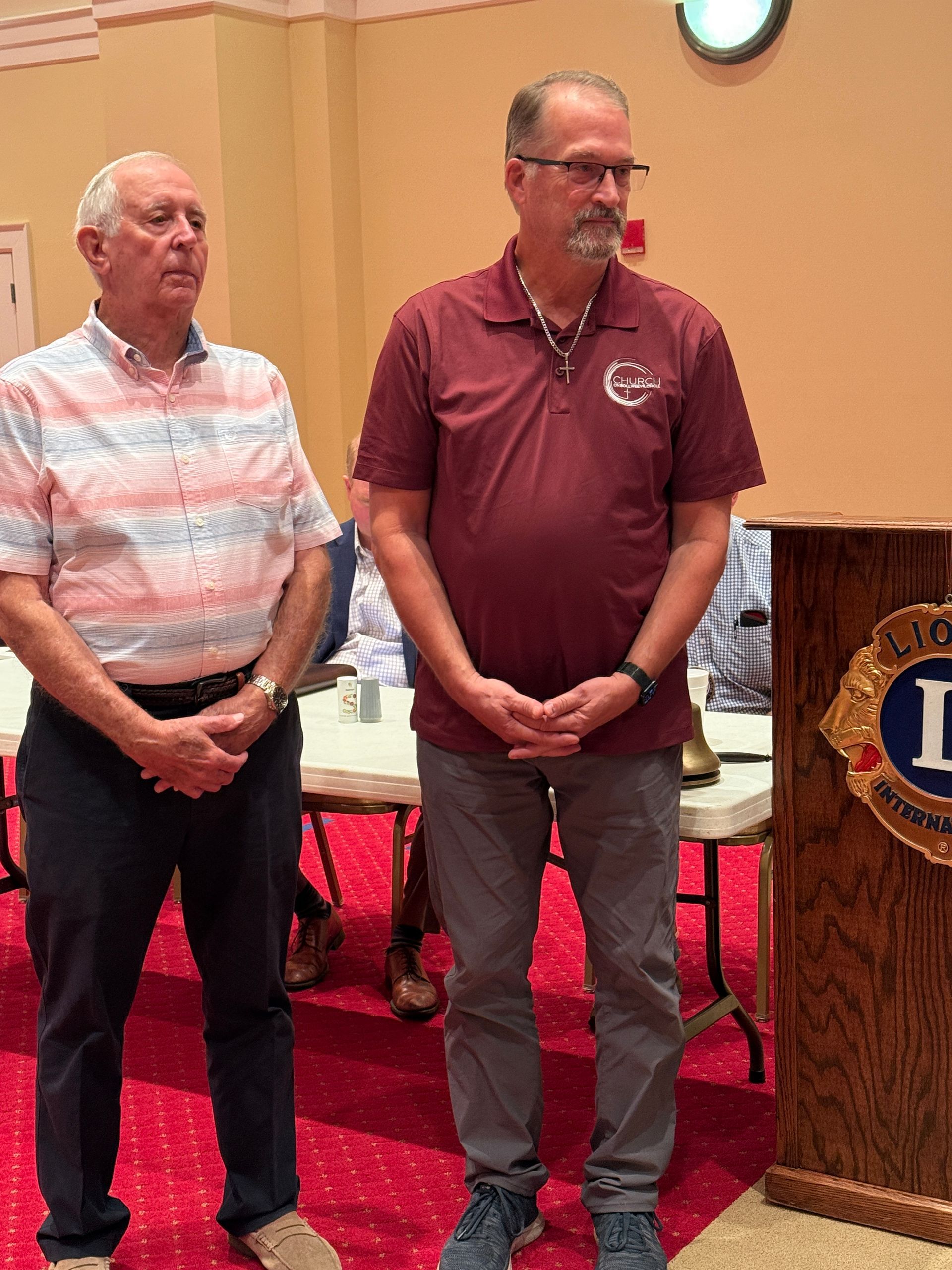 Two men standing by a podium in a room. One in a red shirt, the other in a patterned shirt.