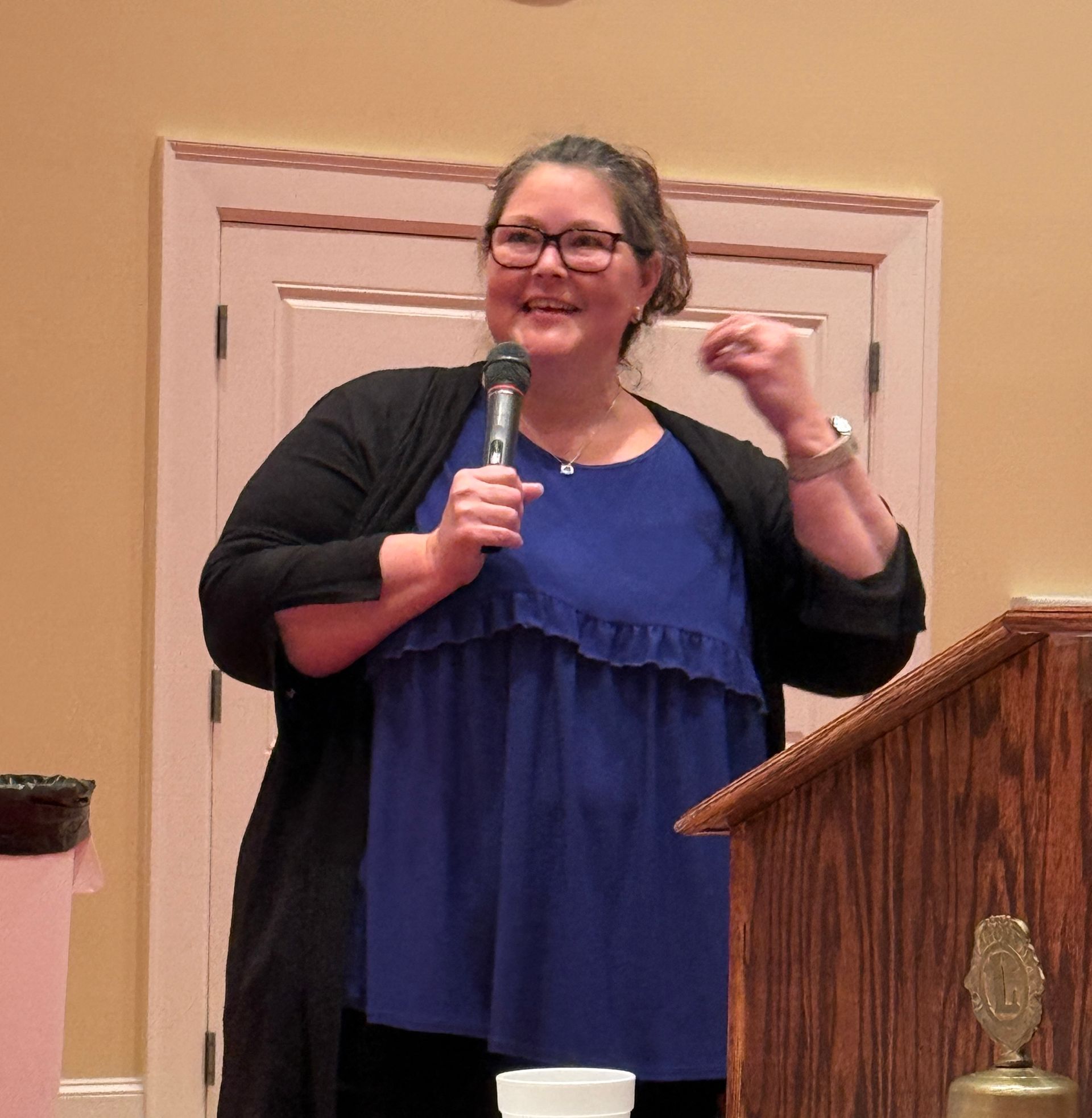Woman speaking at a podium, holding a microphone. Wearing blue top, black cardigan, glasses, and smiling.