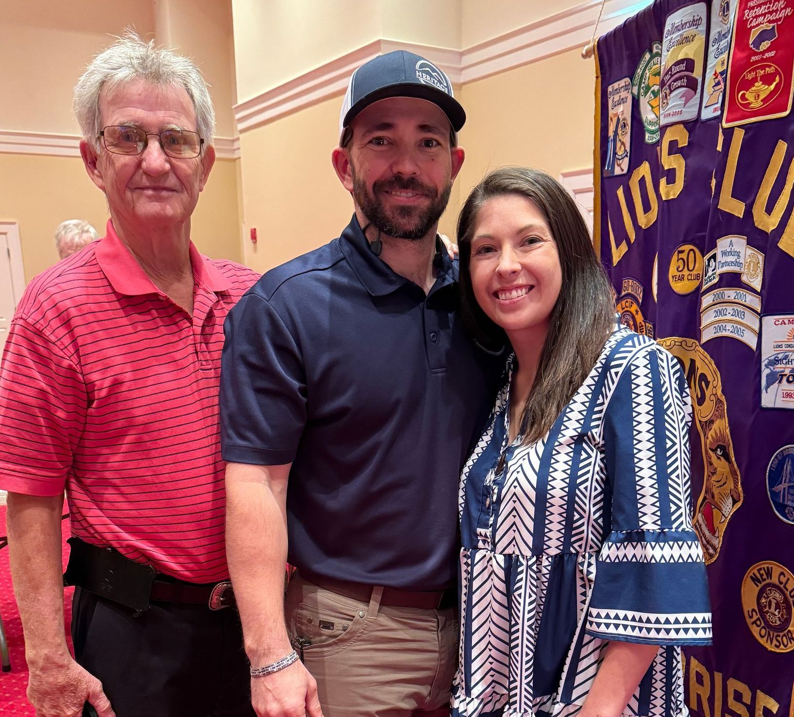Three people smiling in front of a Lions Club banner. Man in red shirt, man in blue shirt/hat, woman in blue and white dress.