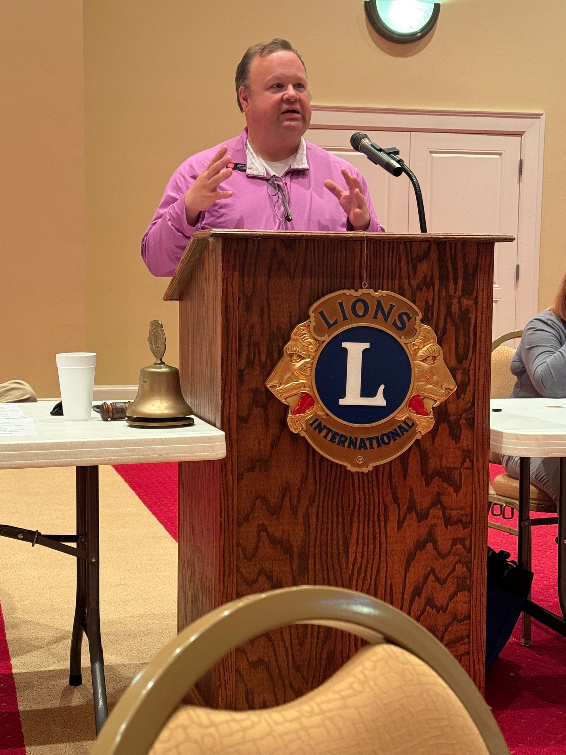 Man speaking at a Lions Club podium, wearing a pink shirt. The podium has the Lions International logo.