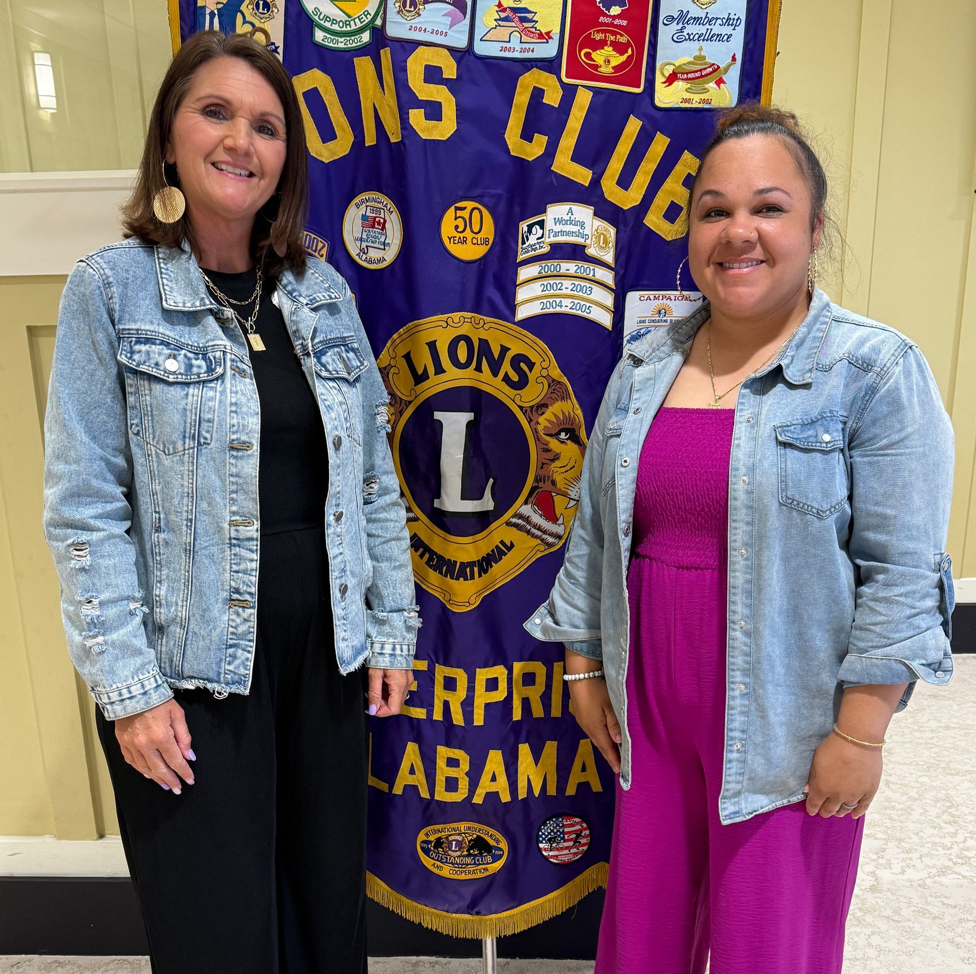 Two women standing beside a Lions Club banner in a hallway, both wearing denim jackets.