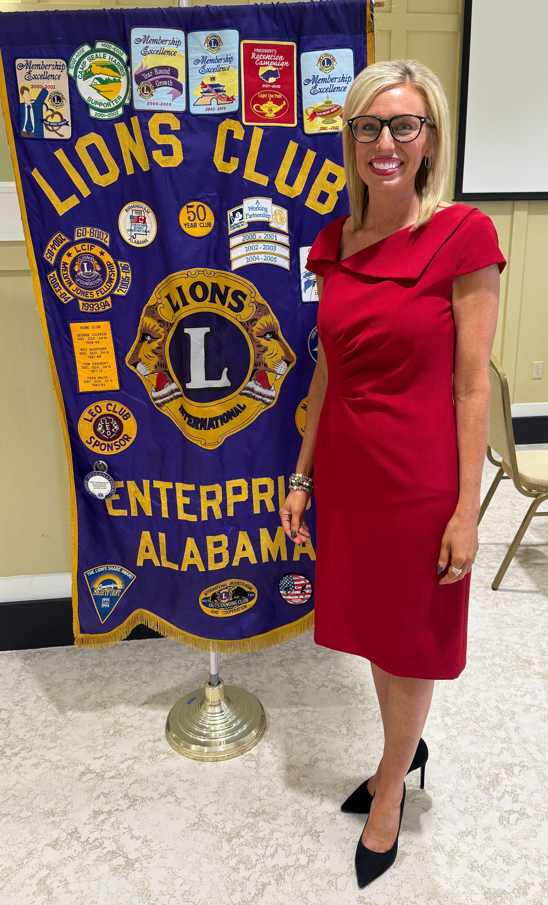 Woman in red dress standing beside a Lions Club banner at an indoor event