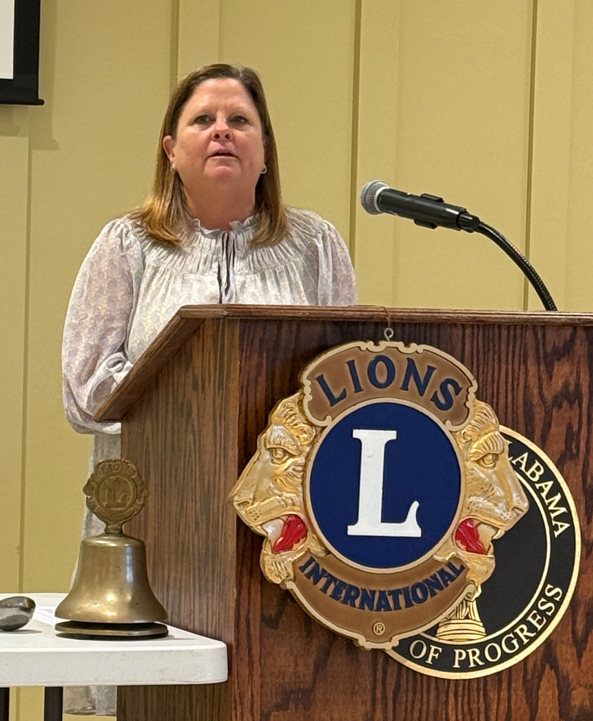Woman speaking at a podium with a Lions International emblem and microphone beside a bell