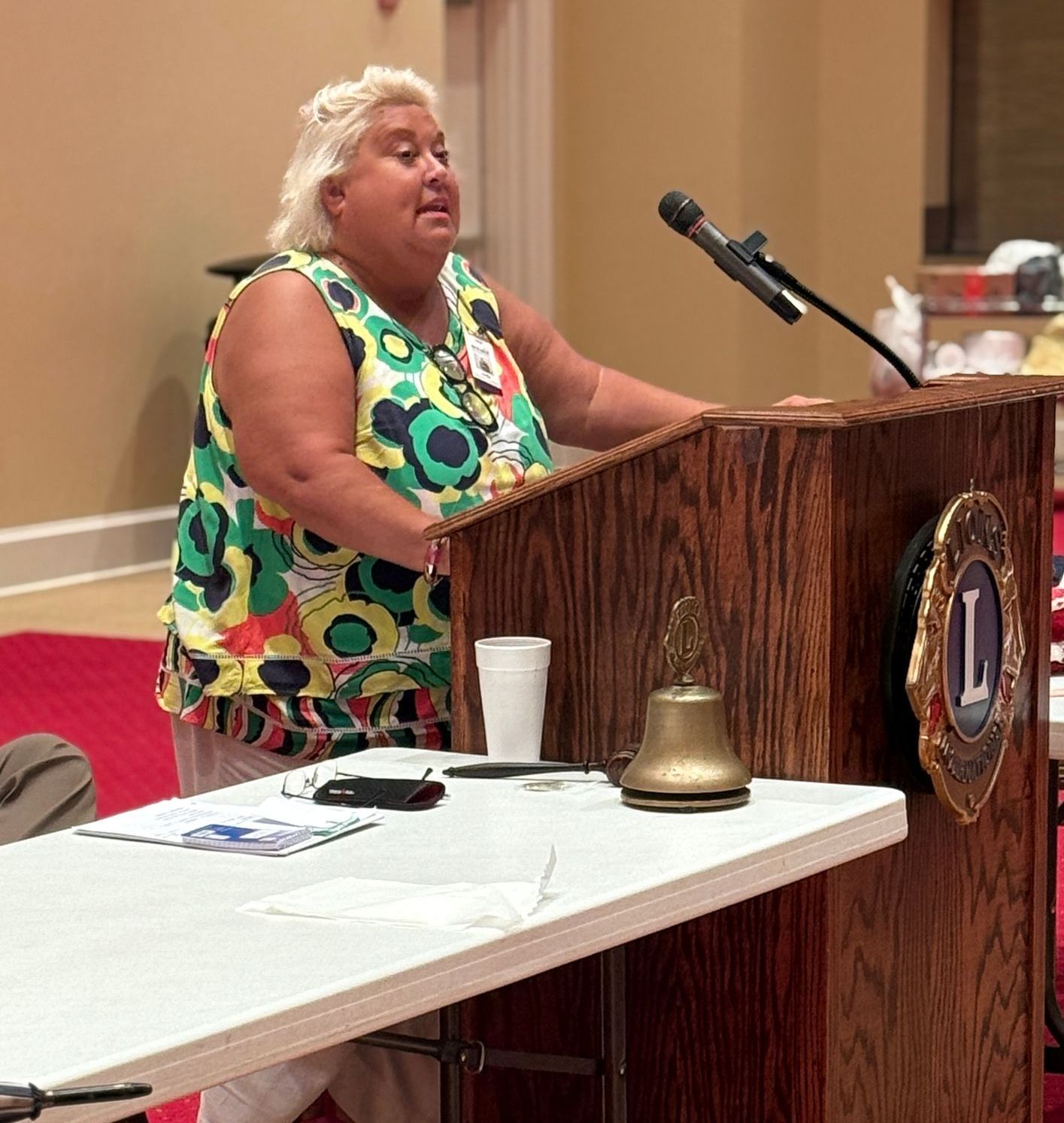 Woman speaking at a podium; colorful shirt; Lions Club emblem.