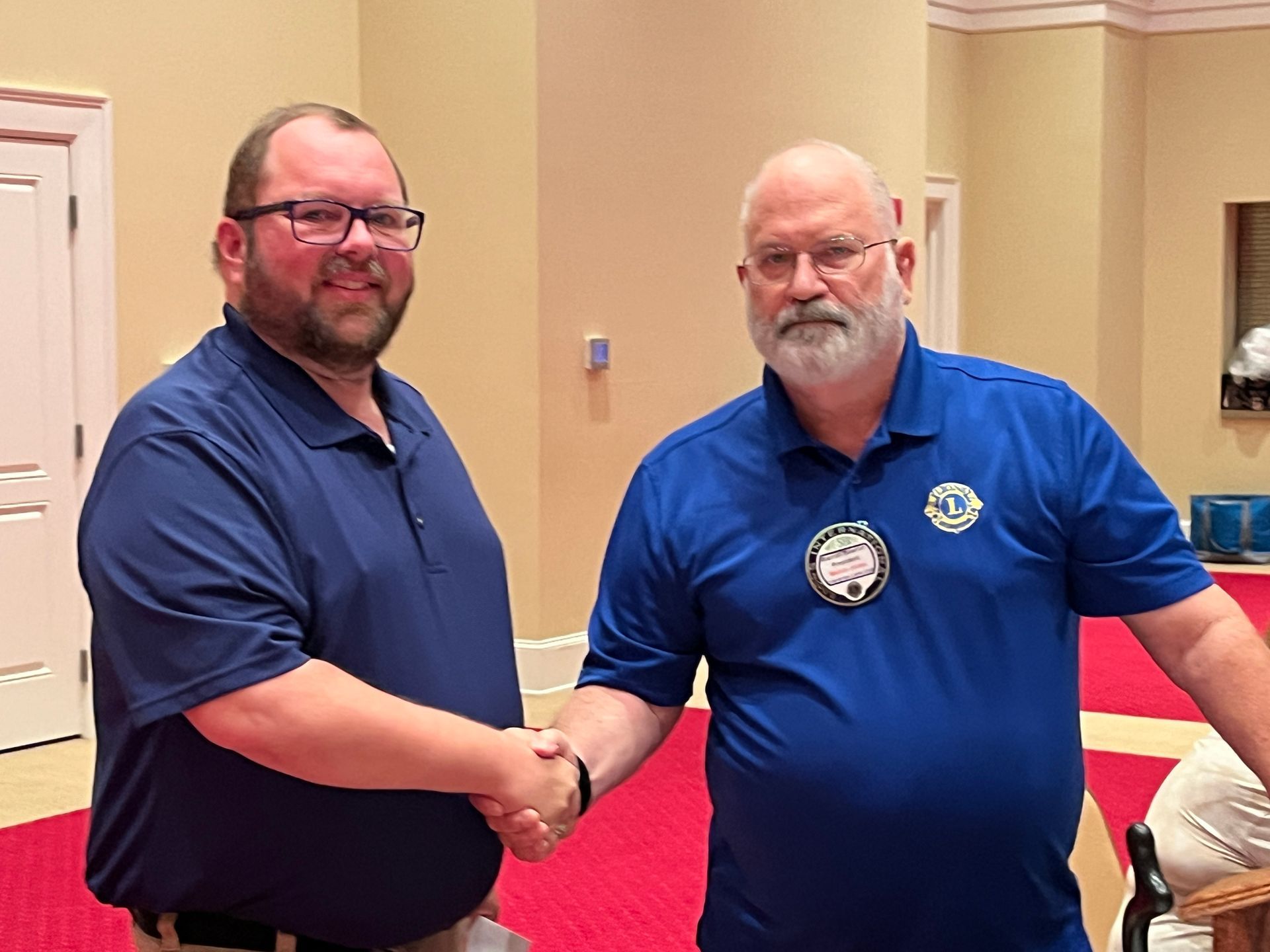 Two men shaking hands, both wearing blue shirts, indoors.