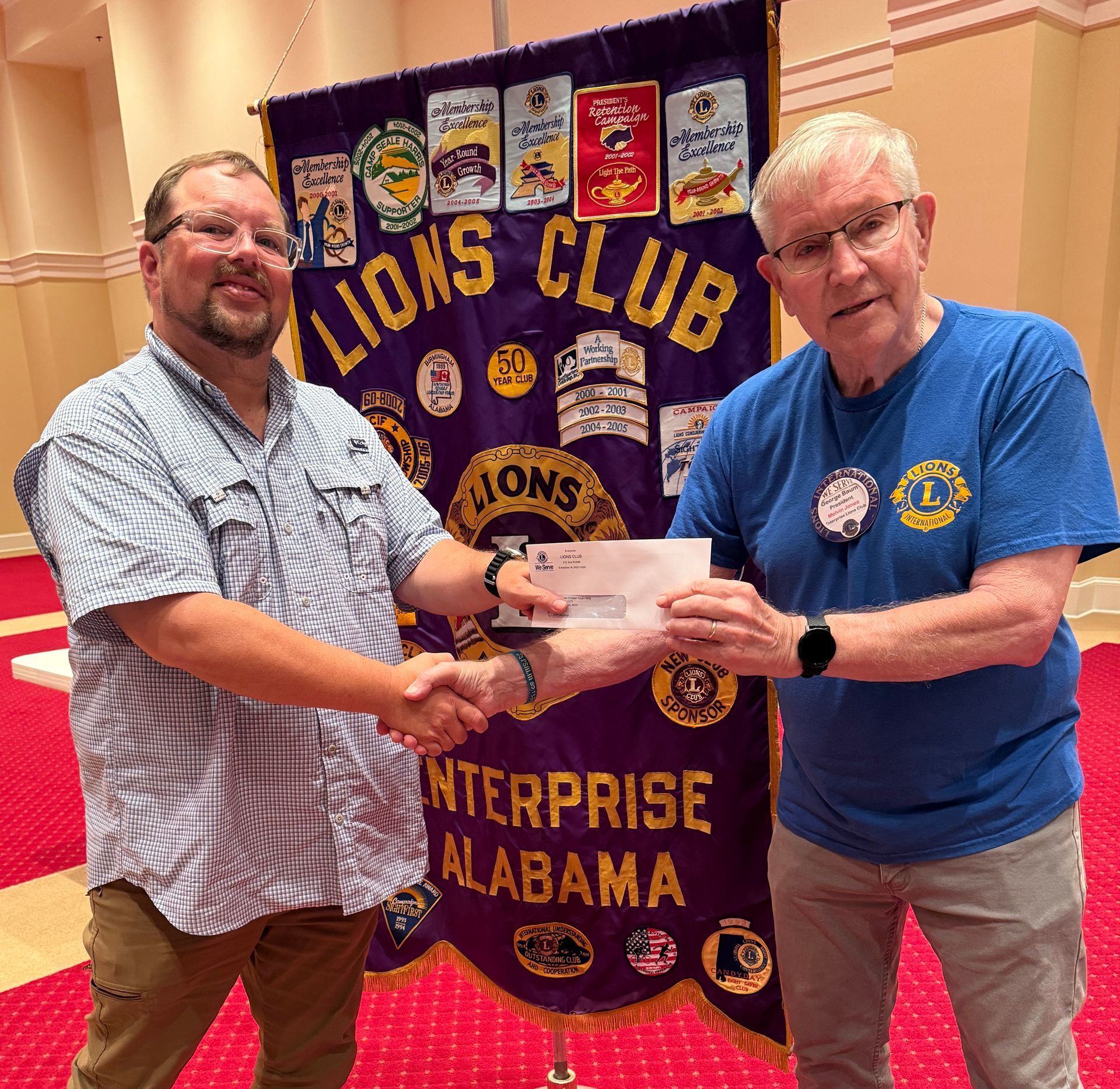 Two men shaking hands, one giving a check in front of a Lions Club banner.