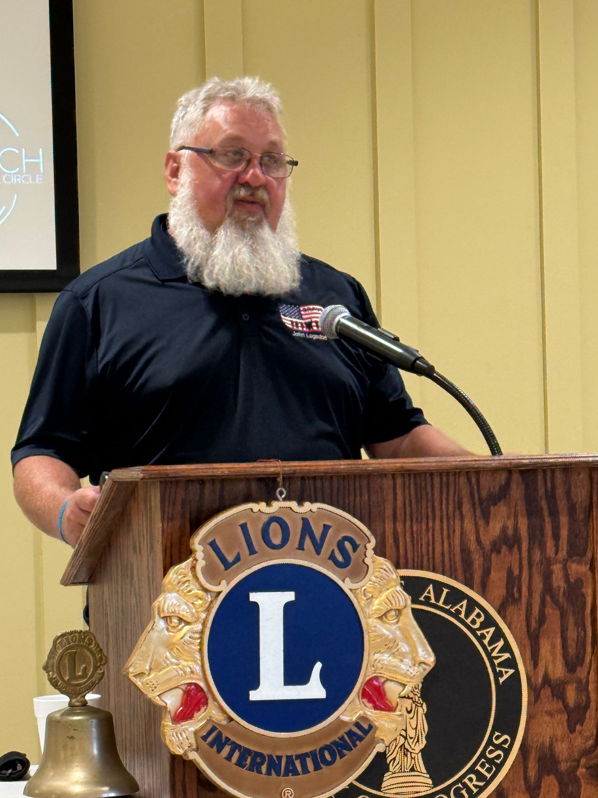Man speaking at a Lions International podium with a microphone and gold Lions emblem.