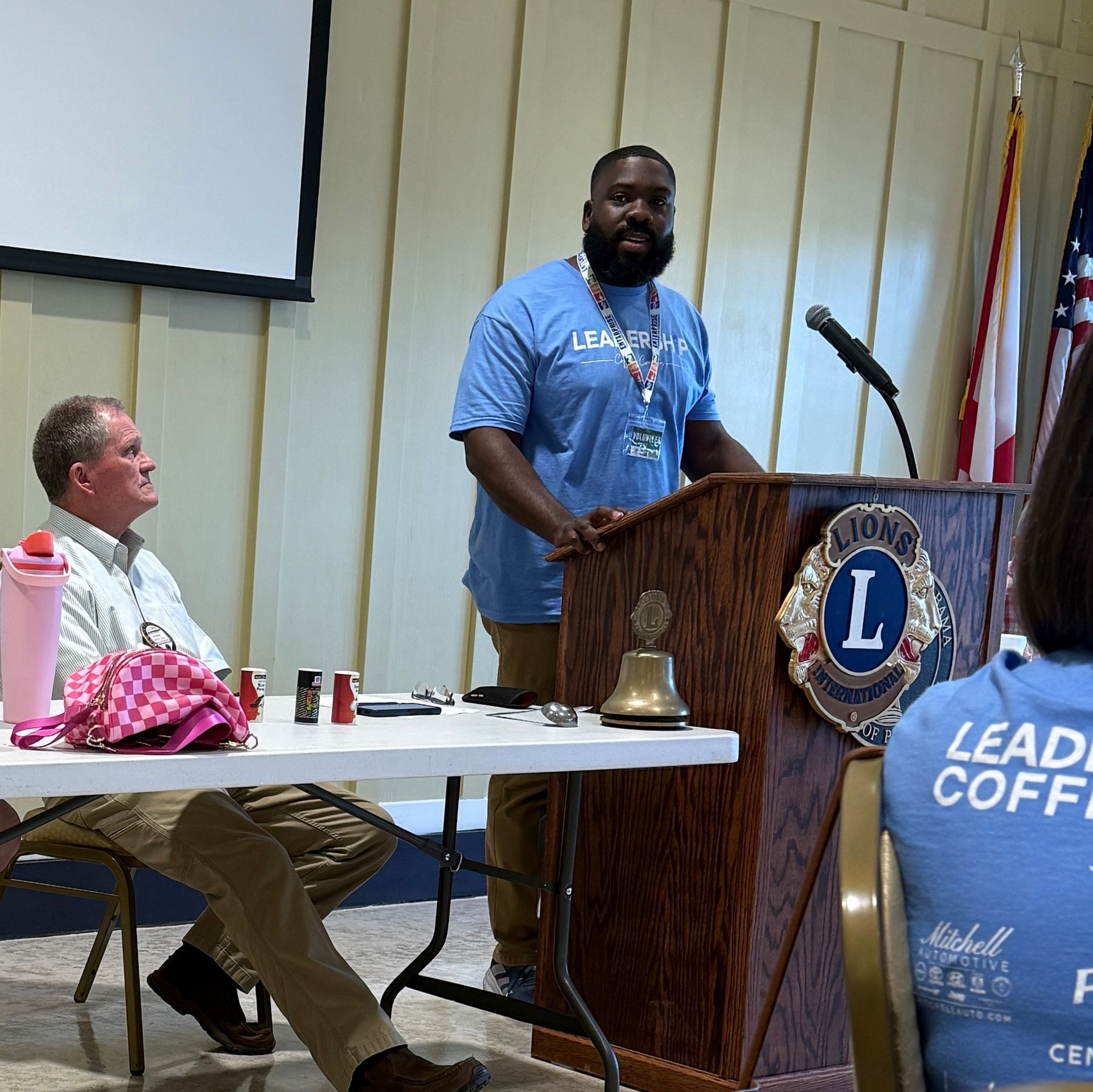 Man speaking at a podium during a Lions Club meeting, with seated audience and a projection screen nearby