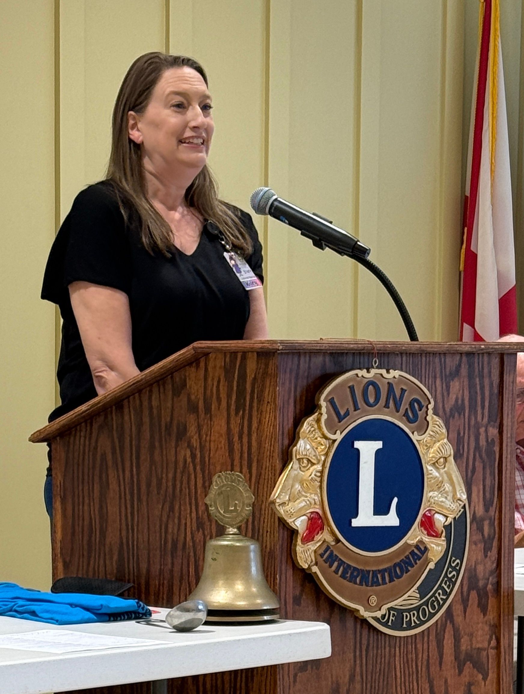 Woman speaking at a podium with a Lions Club emblem, microphone, and bell in a meeting room