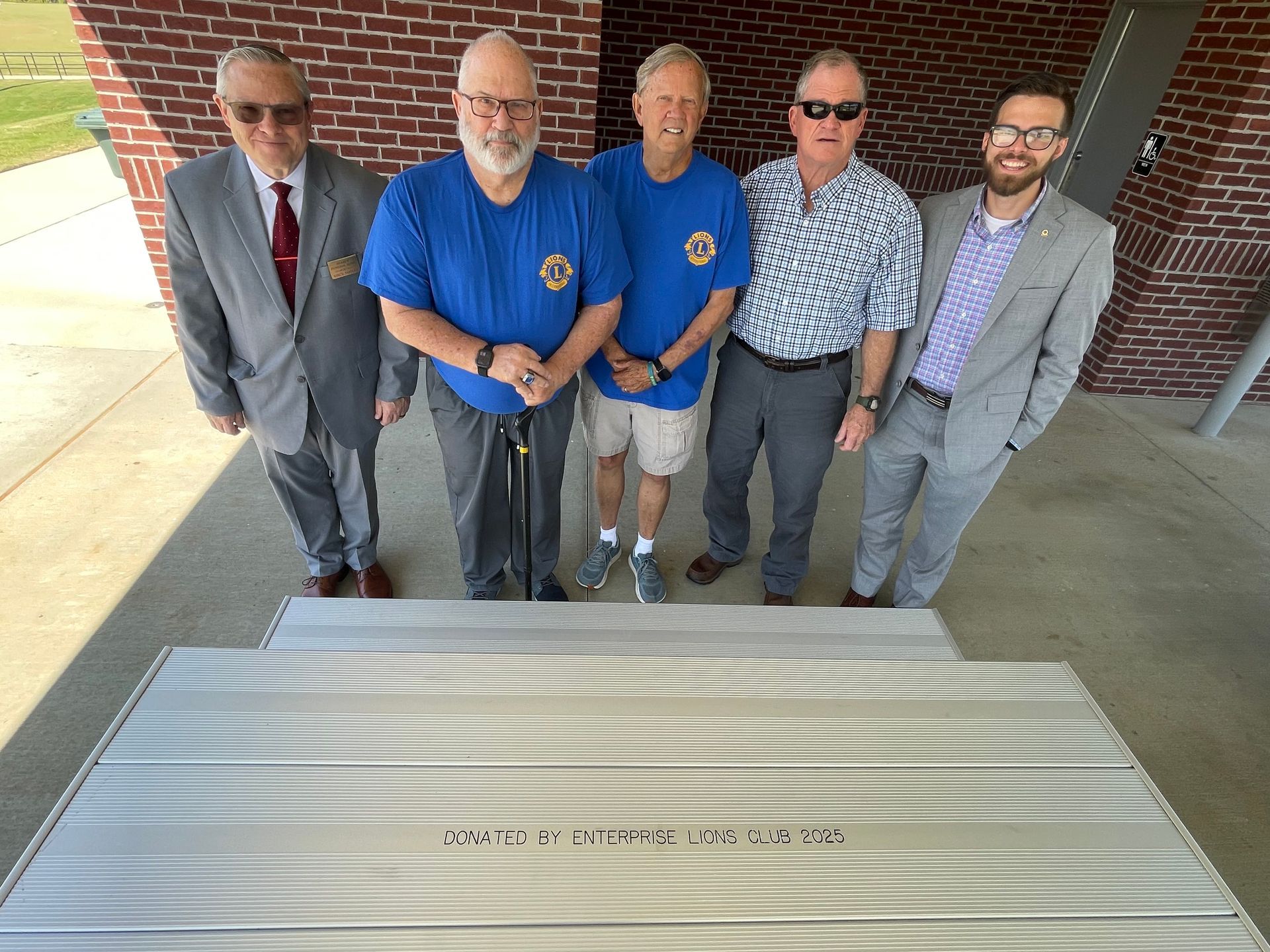 Five men stand behind a large, metallic plaque outdoors. They are smiling, and two wear blue shirts.