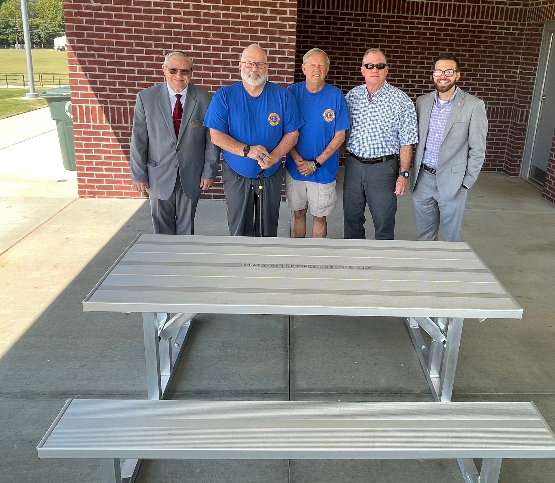 Five men stand near a picnic table under a covered area, a park in background.