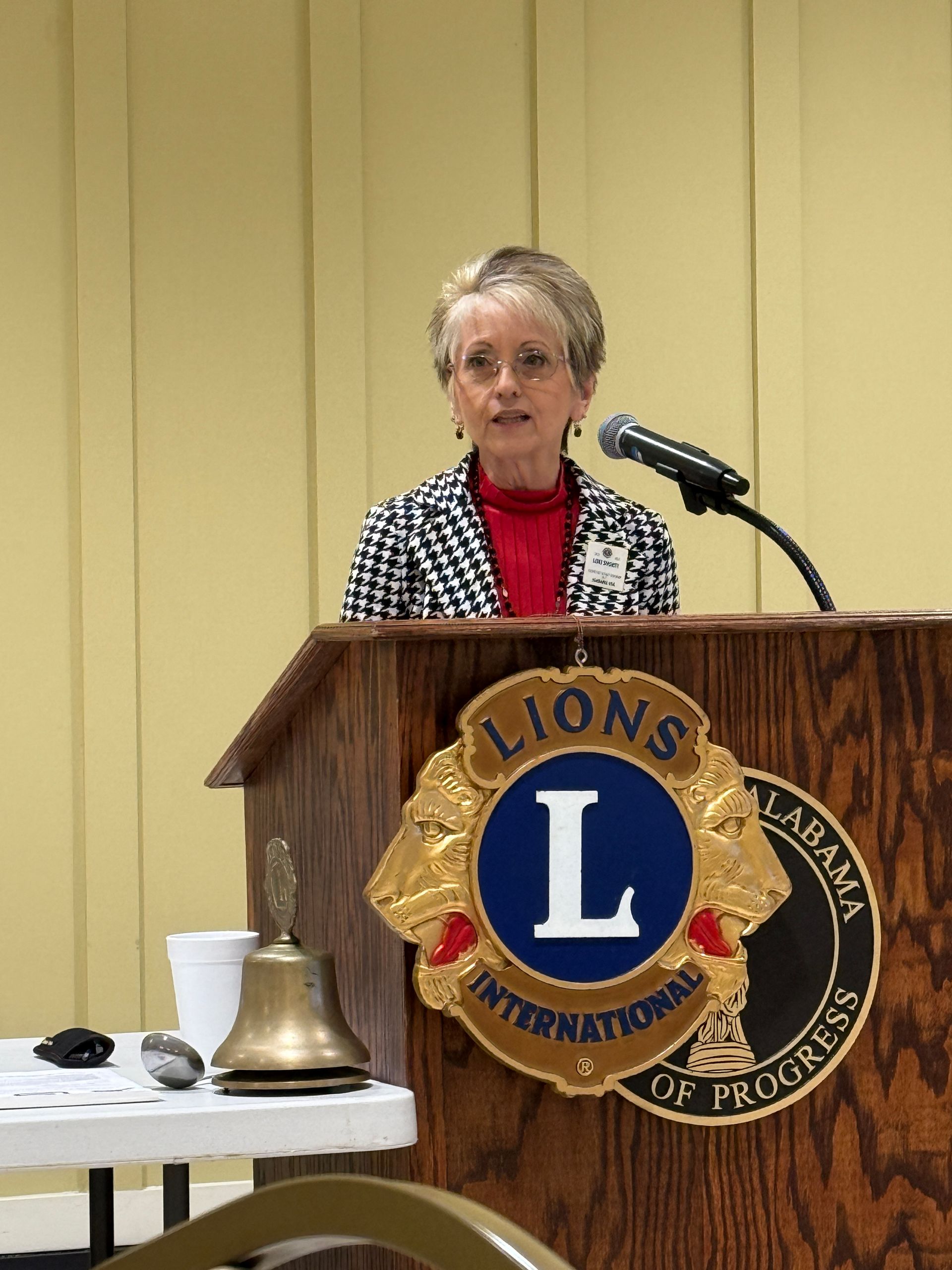 Speaker at a podium with Lions Club emblem, speaking into a microphone at a meeting