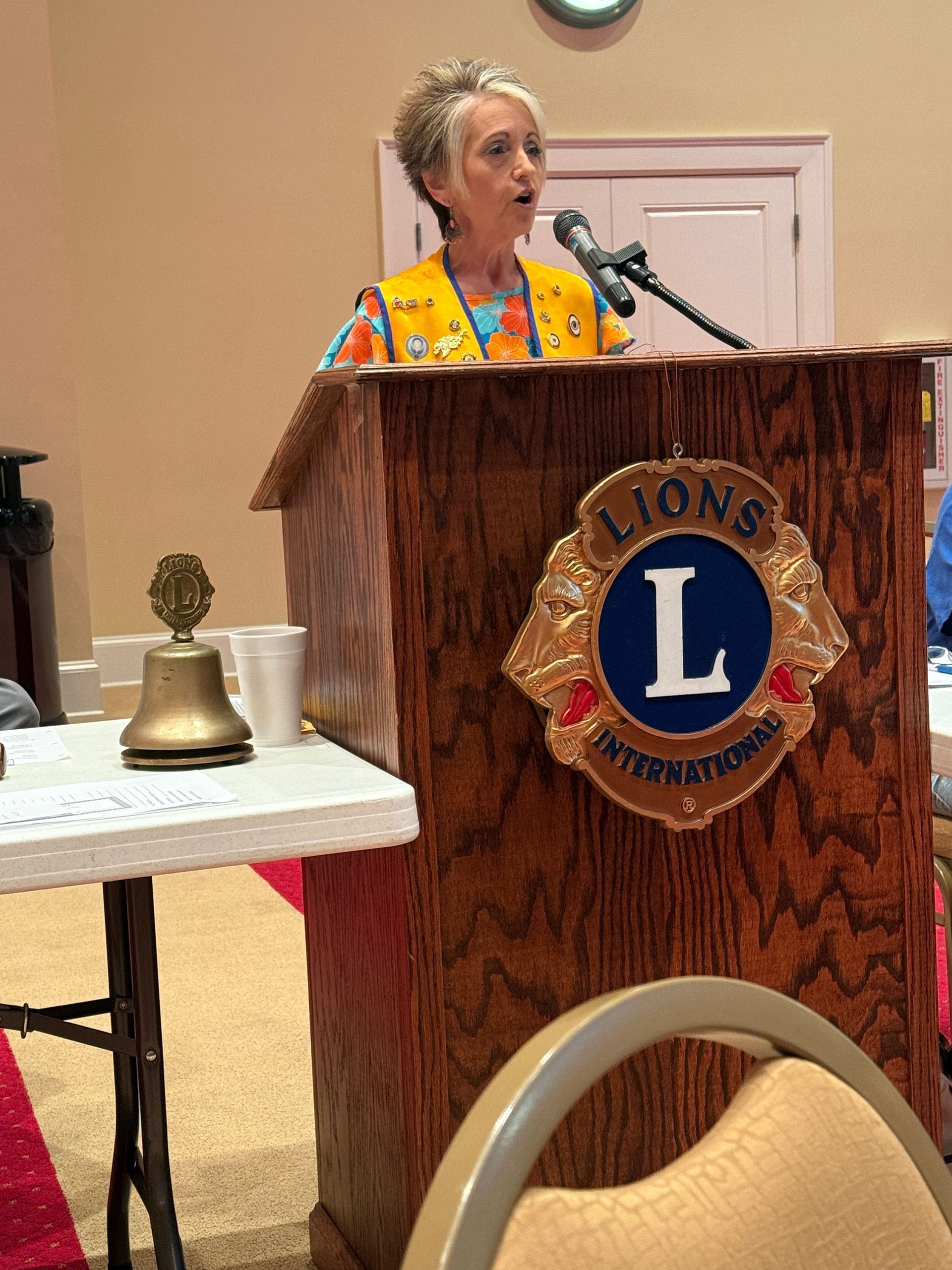 Woman speaking at a Lions Club podium, wearing a vest. Wooden podium with Lions International logo.