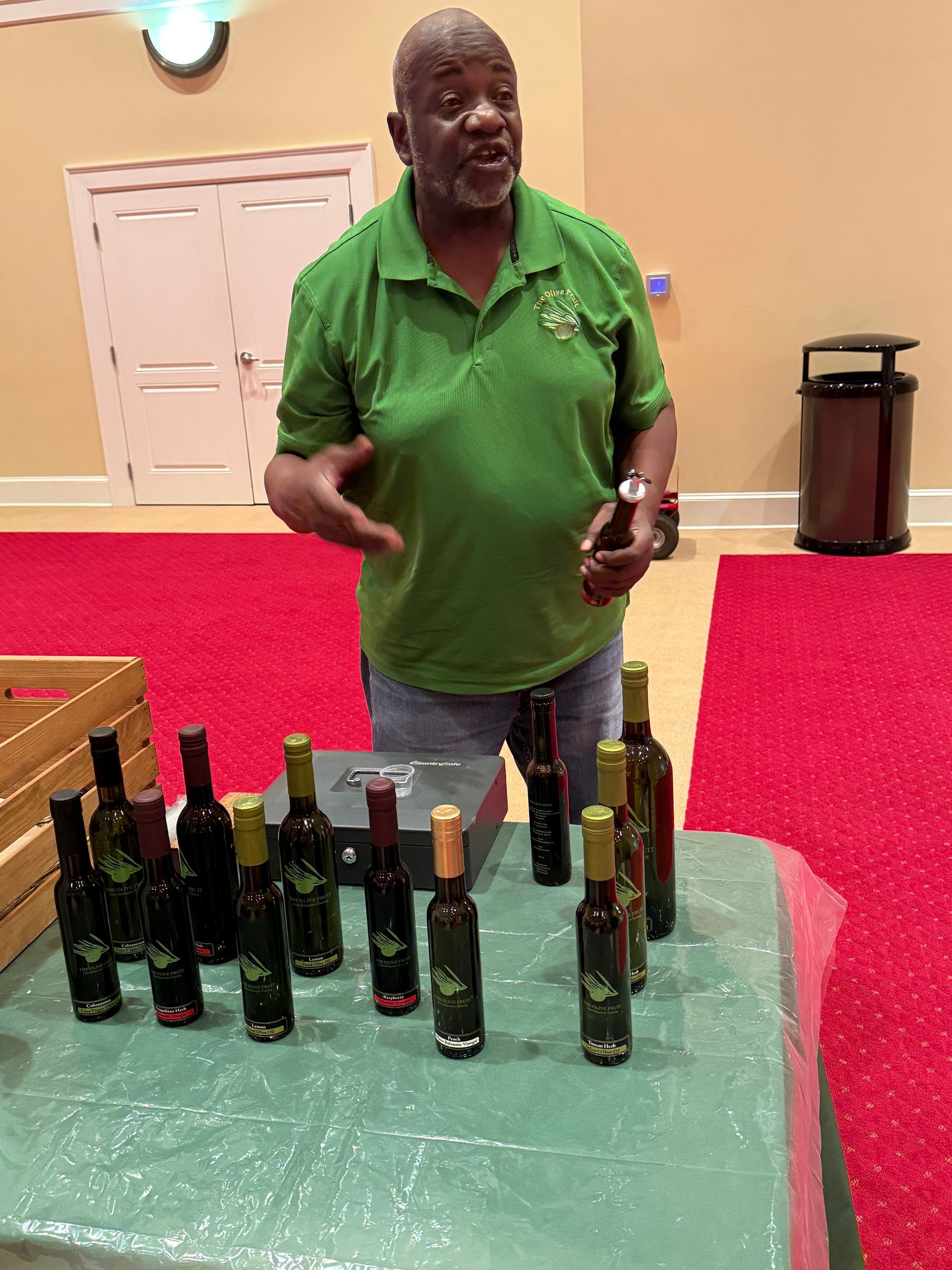Man in green shirt presents olive oil bottles on a table at an event.