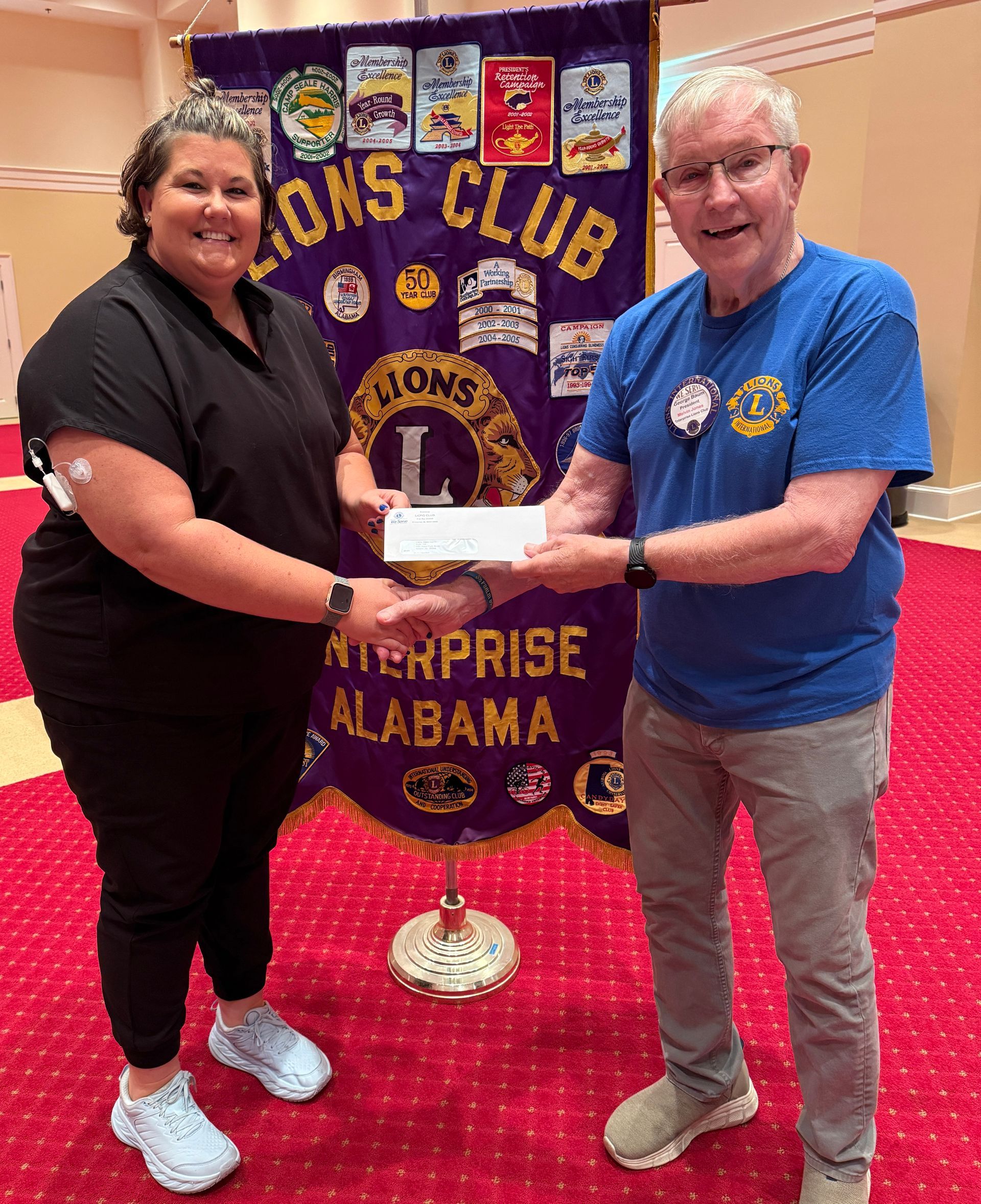 Woman in black shirt receiving a check from a man in a blue shirt, Lions Club banner in background.