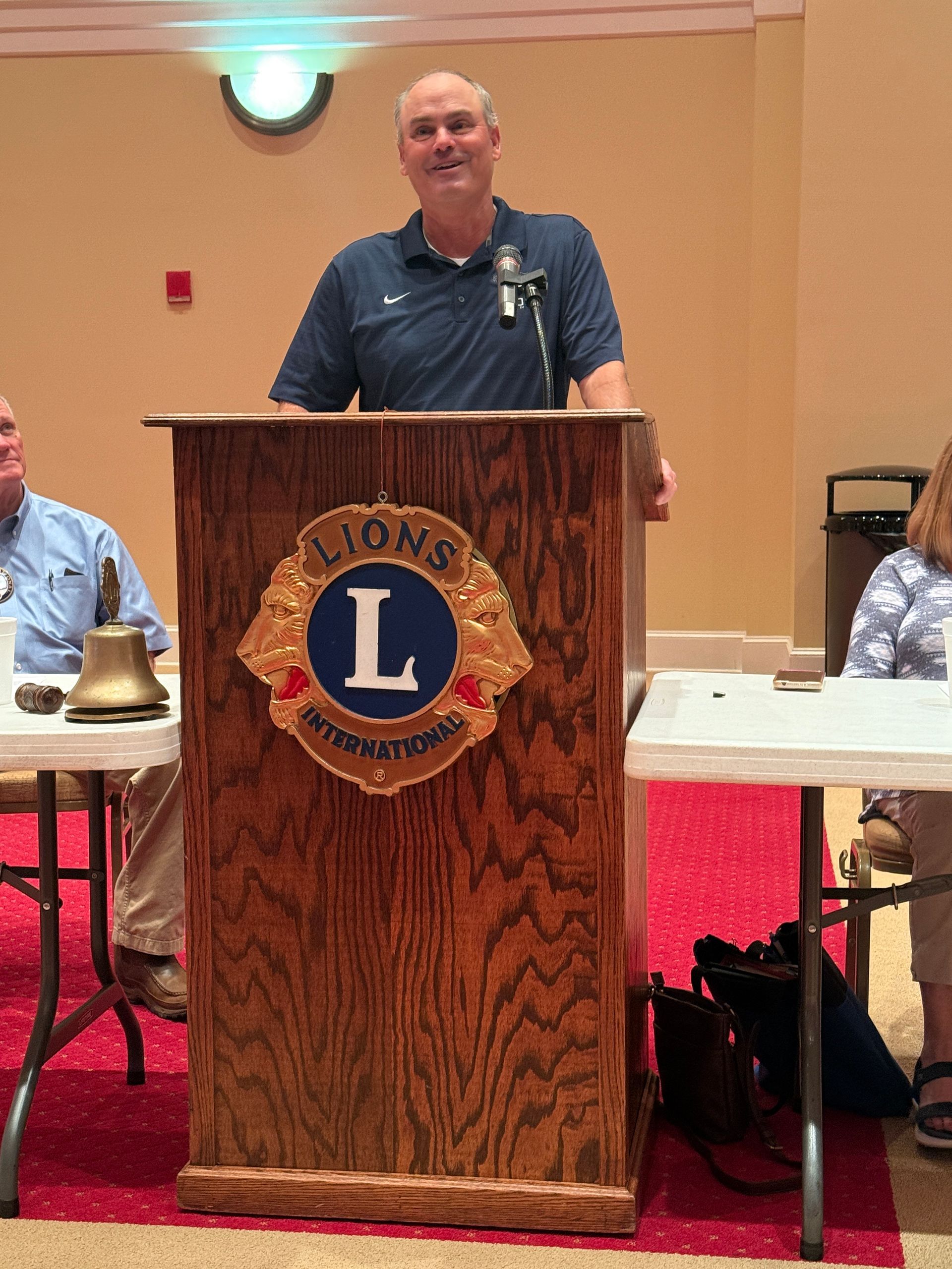 Man speaking at a Lions Club podium. He wears a navy shirt.  Lions emblem on the podium.  Room setting.