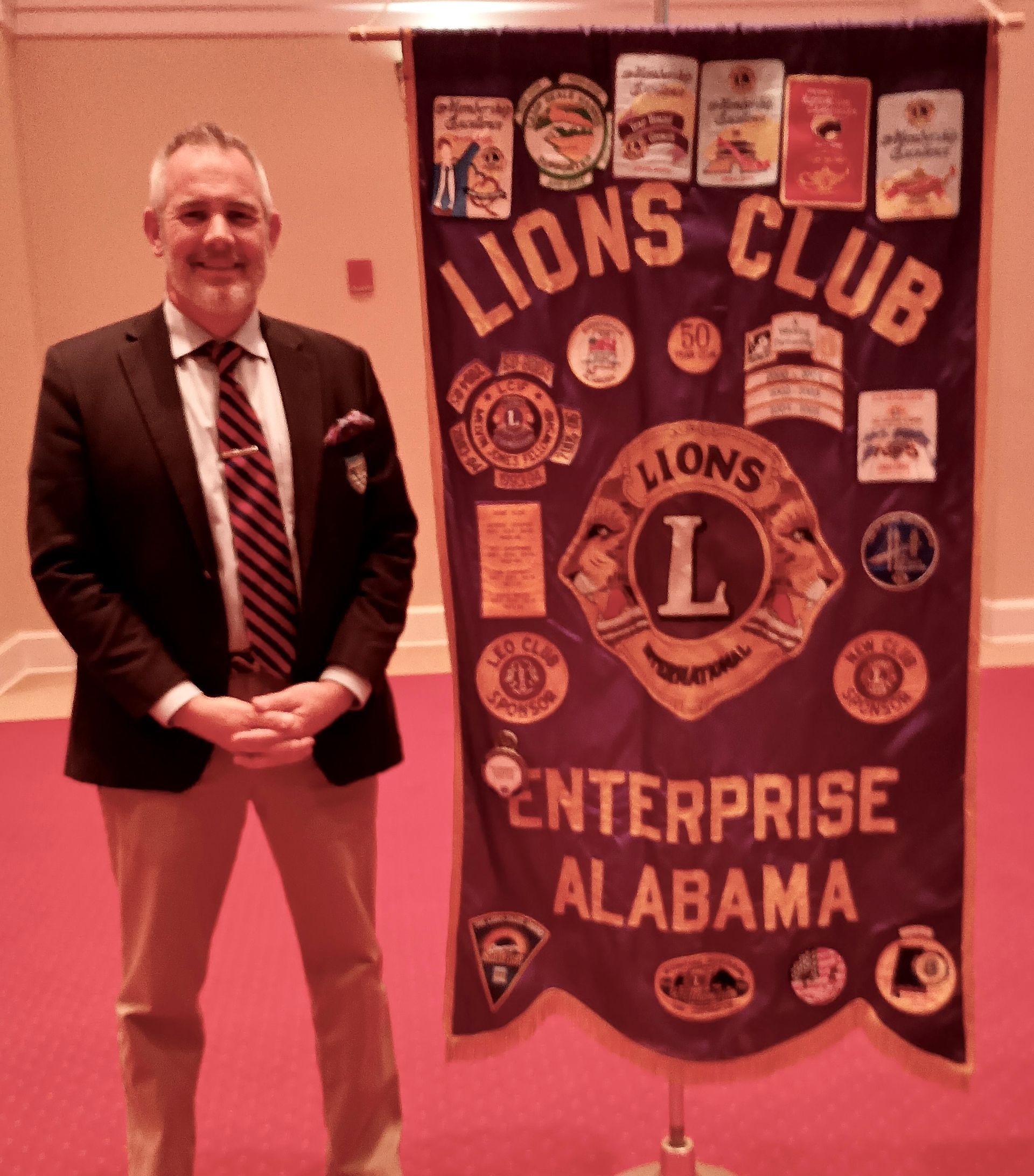 Man in suit stands beside a Lions Club banner; 
