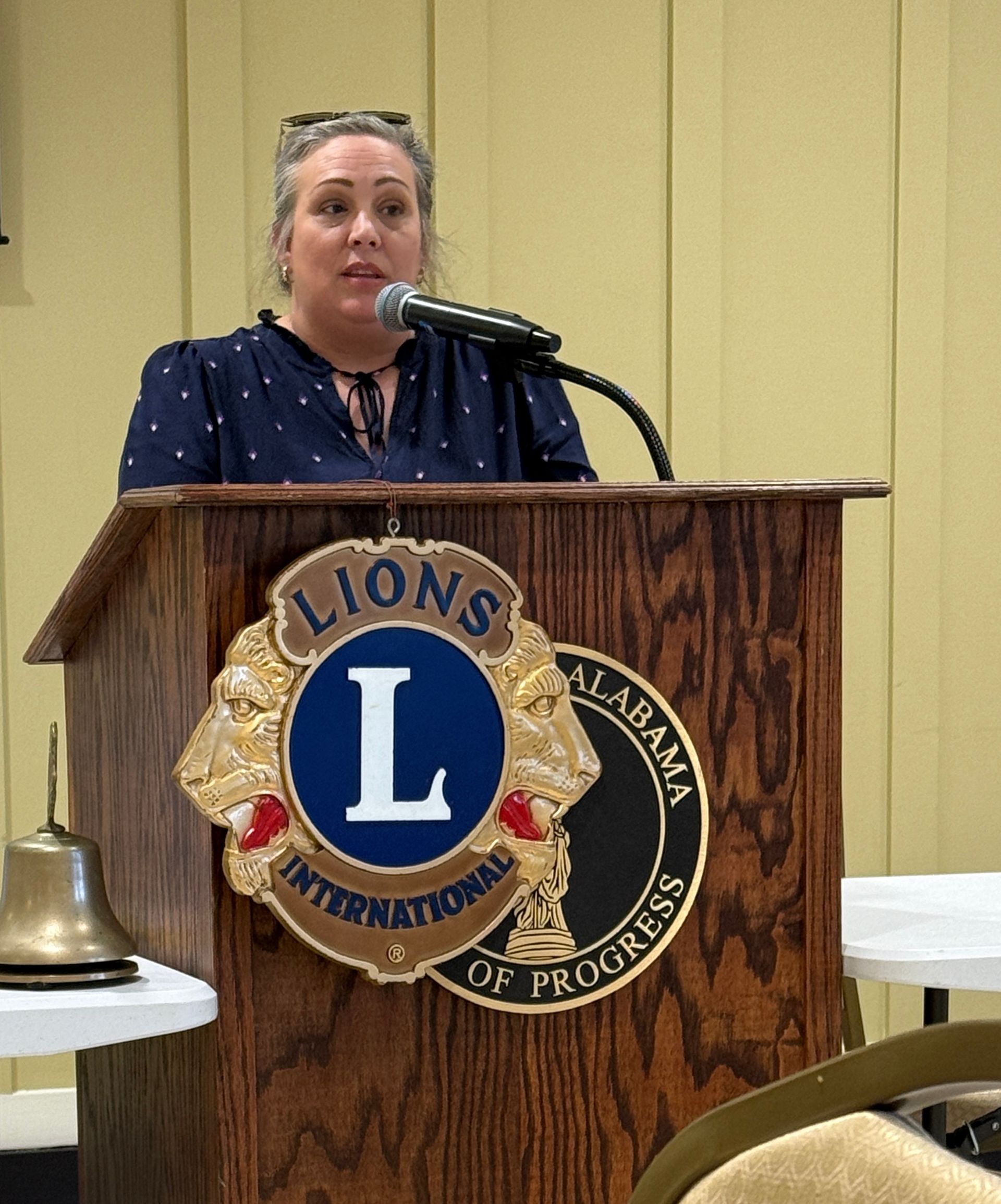 Woman speaking at a podium with Lions Club emblem in a meeting hall