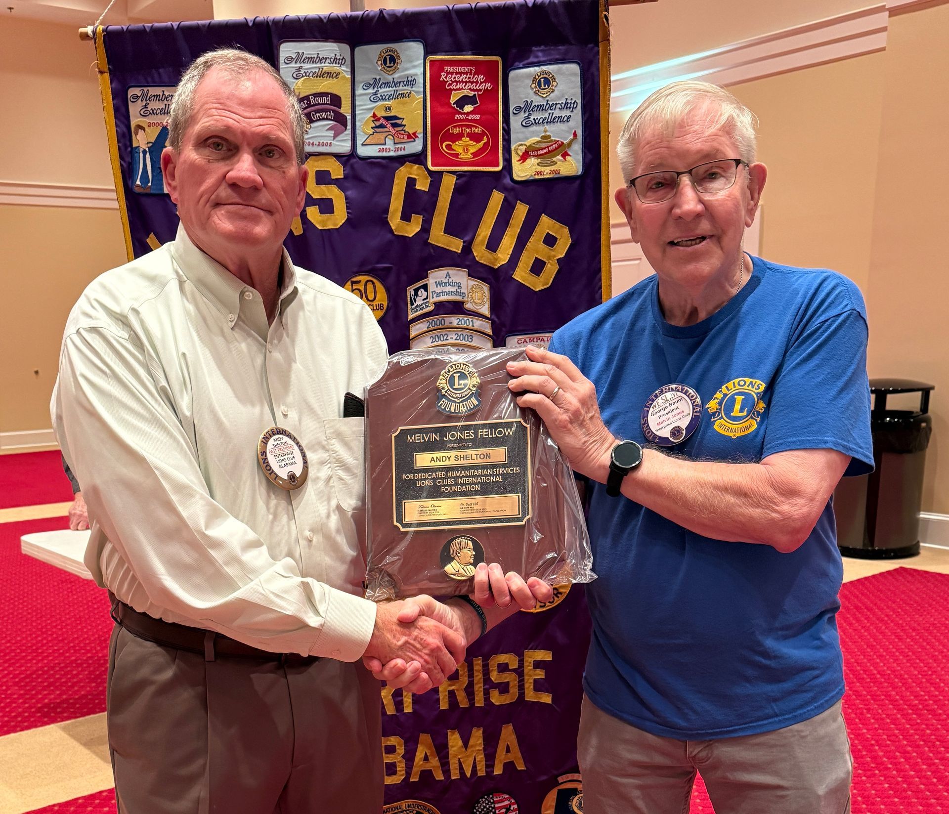 Two men shaking hands, one holding a plaque, in front of a Lions Club banner; indoors with red carpet.