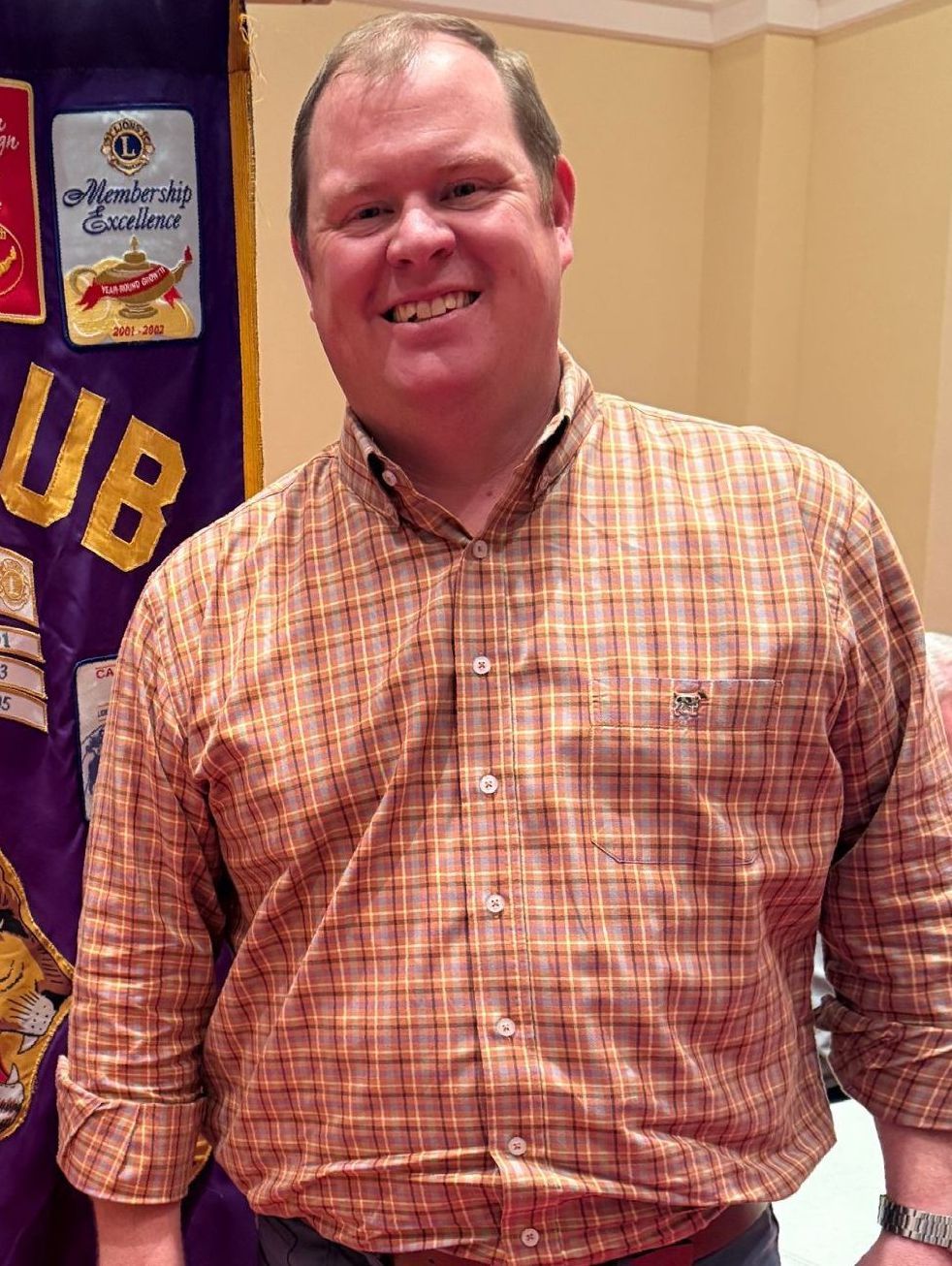 Man in plaid shirt smiles, standing in front of a purple Lions Club banner.