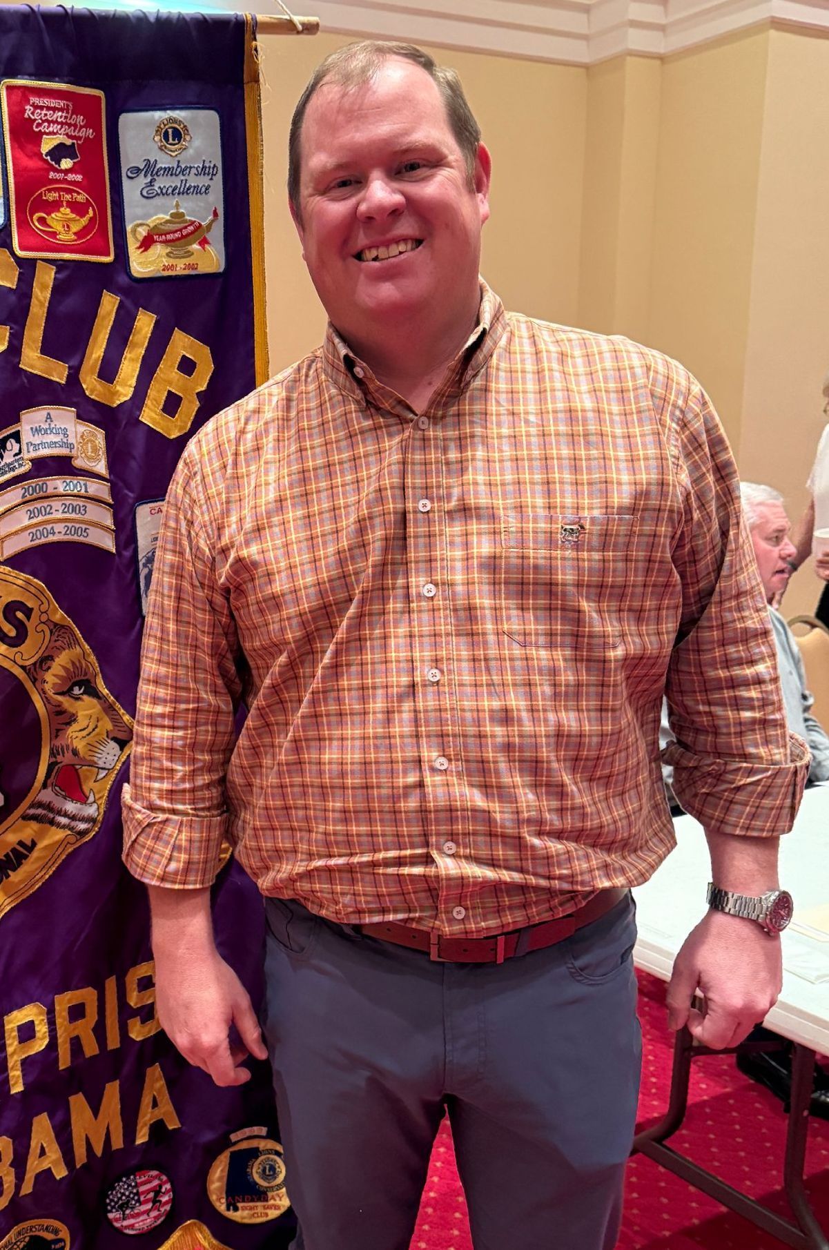 Man in patterned shirt smiles in front of a Lions Club banner; indoor setting.