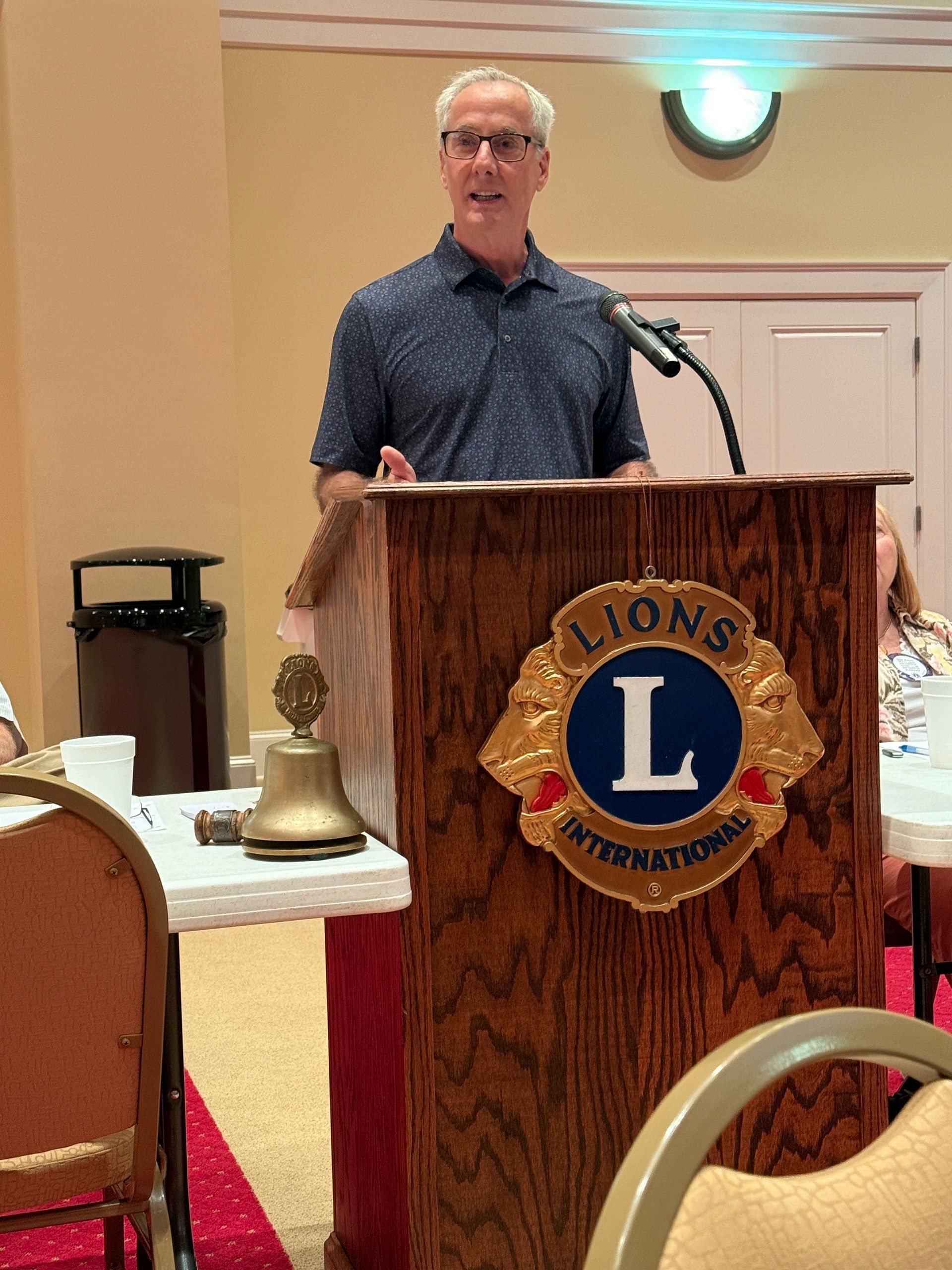 Man speaking at a Lions Club podium, wearing a blue shirt. Red and gold emblem.