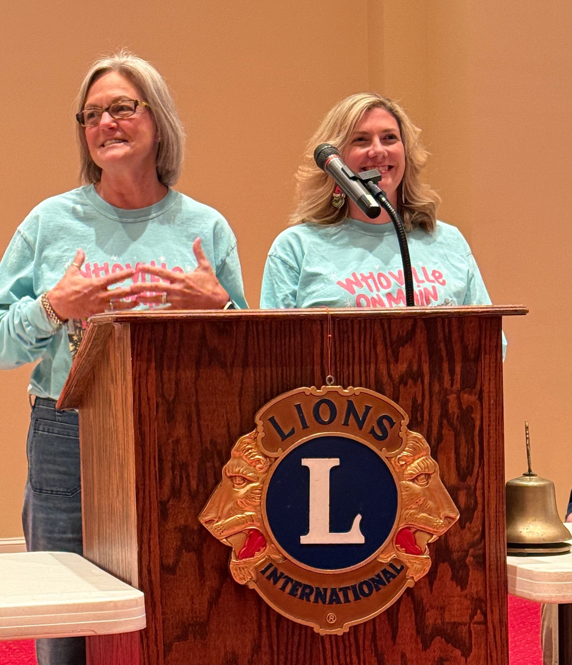 Two women speaking at a Lions Club podium, wearing matching blue shirts. The podium has the Lions International logo.