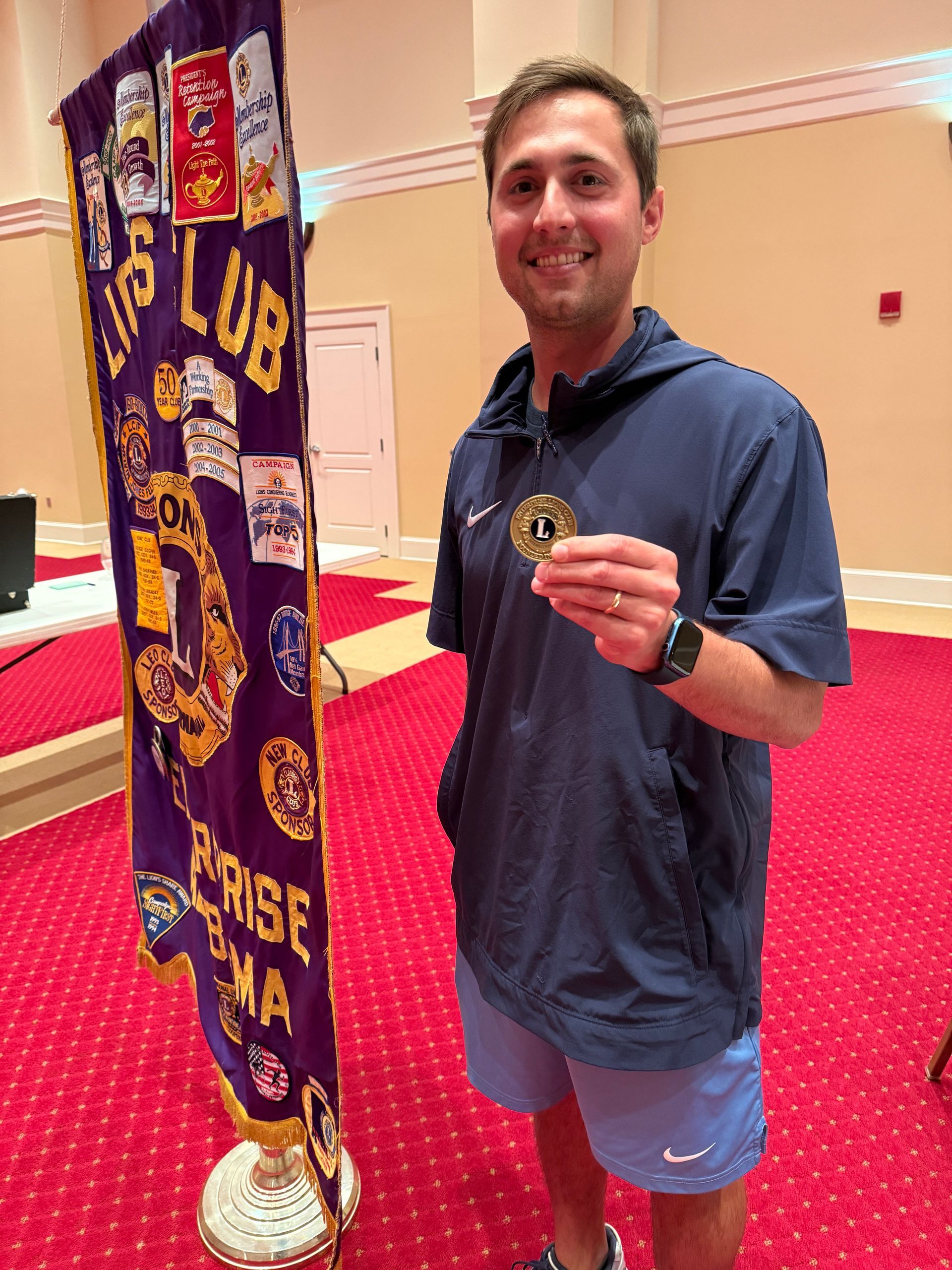 Man smiles, holding a medal, next to a Lions Club banner in a formal setting.
