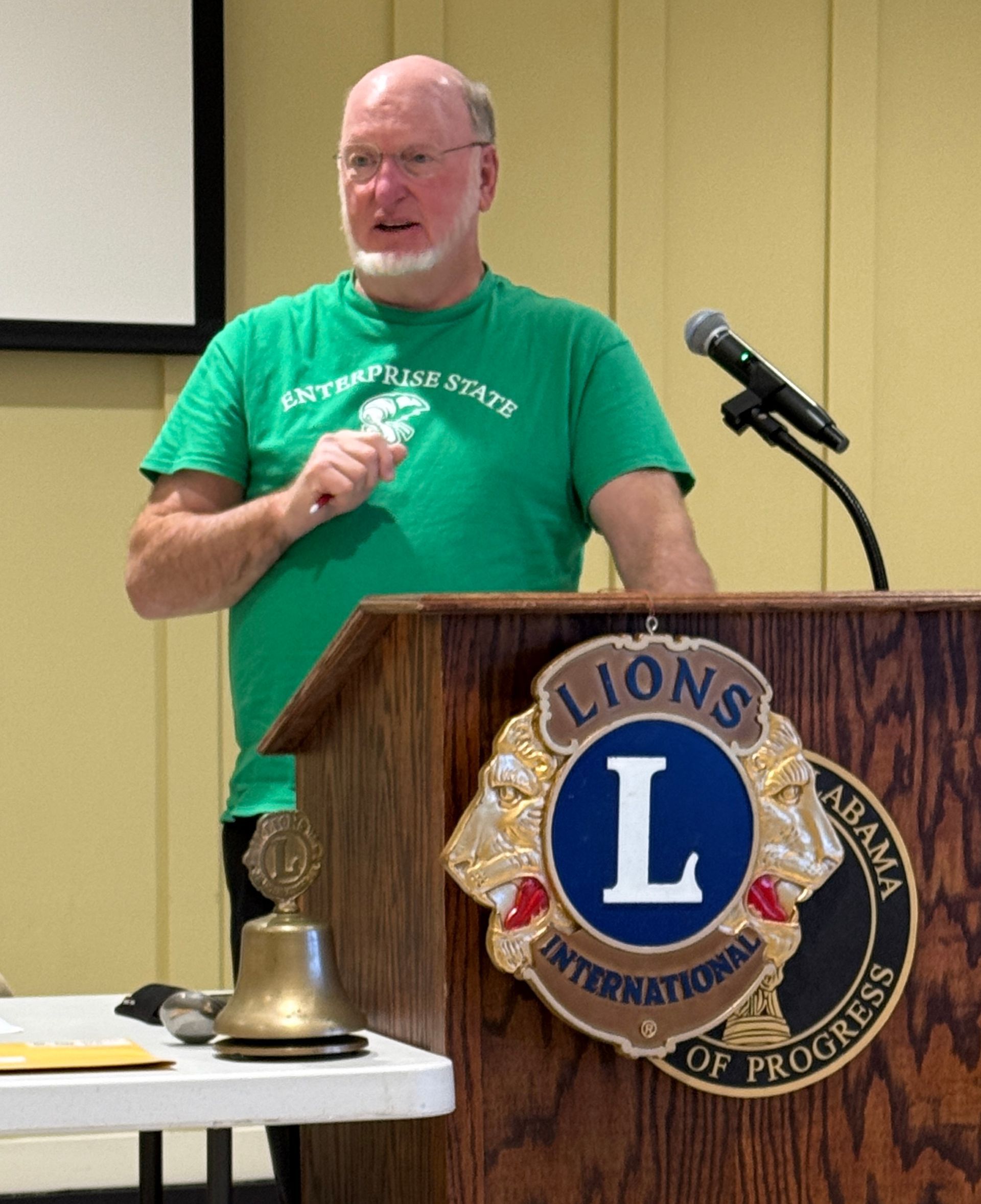 Man speaking at a Lions Club podium in a green T-shirt, with a microphone and brass bell on the table