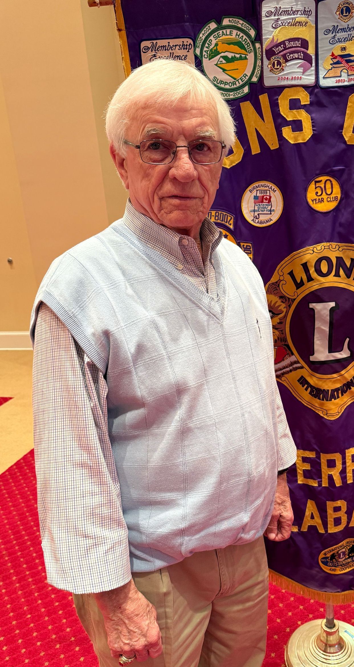 An elderly man with glasses wearing a sweater vest, standing in front of a Lions Club banner.