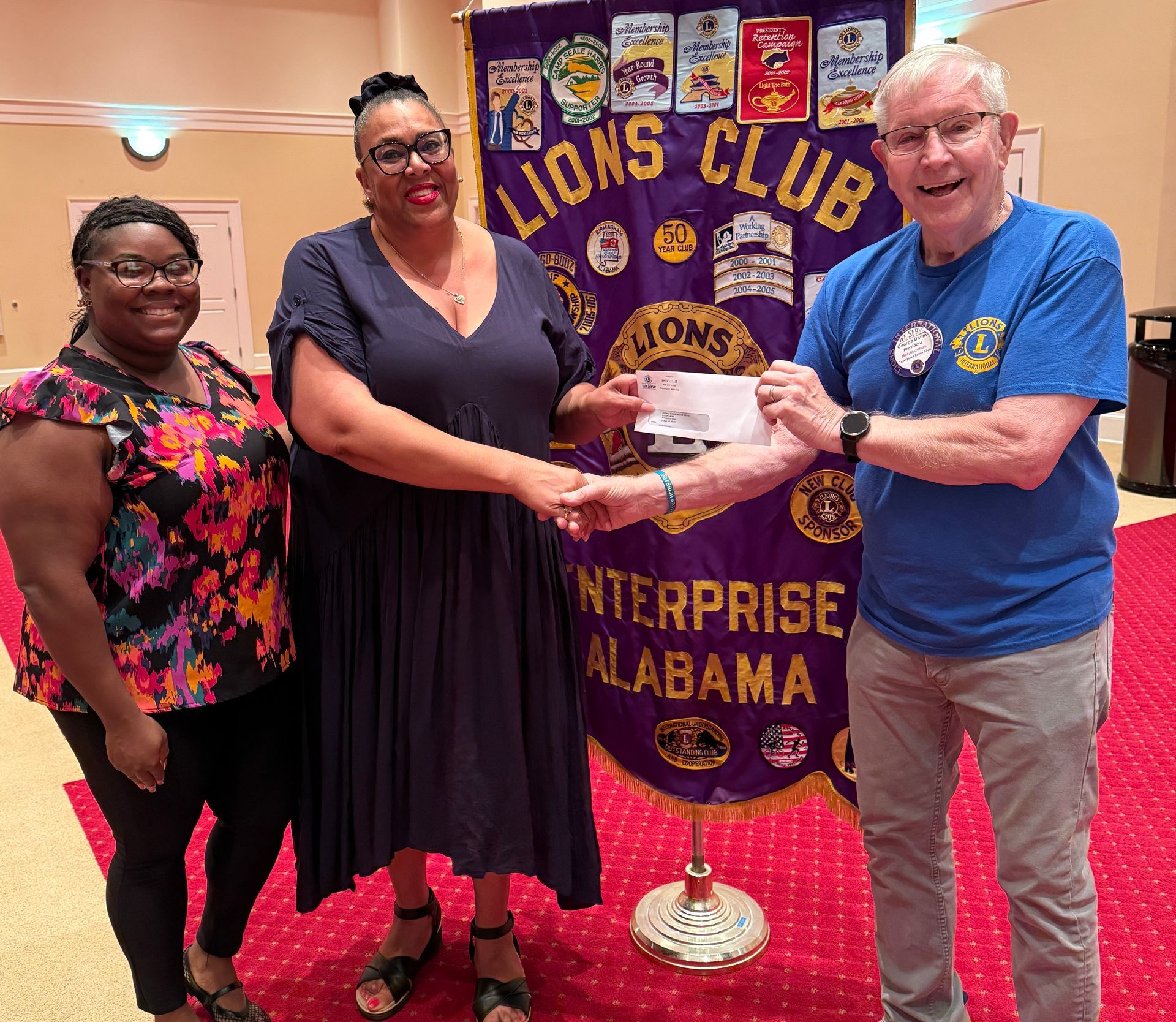 Three people stand in front of a Lions Club banner, one hands a check. Enterprise, Alabama.