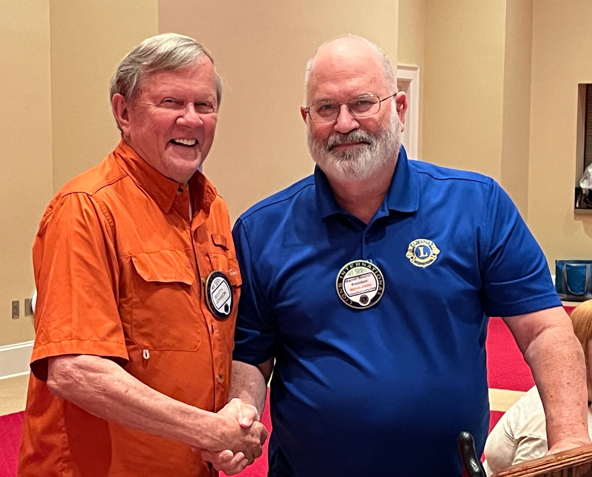 Two men shake hands. One wears orange, one wears blue with a Lions Club pin. Indoor setting.