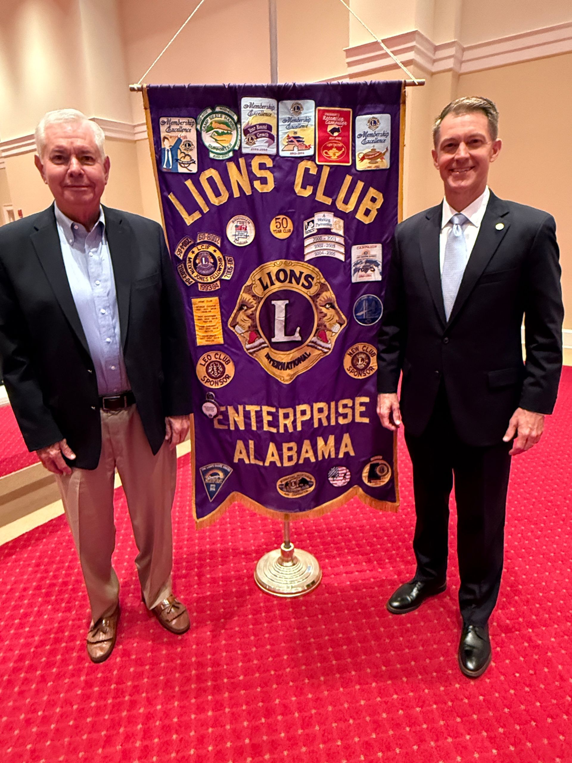 Two men stand by a Lions Club banner in Enterprise, Alabama. One man wears a blazer, the other, a suit.