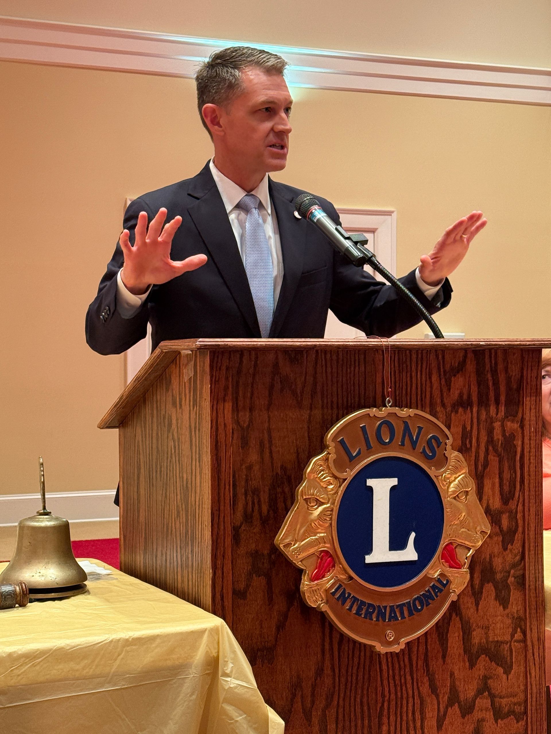 Man speaking at a podium with a Lions International logo; he gestures with hands raised.