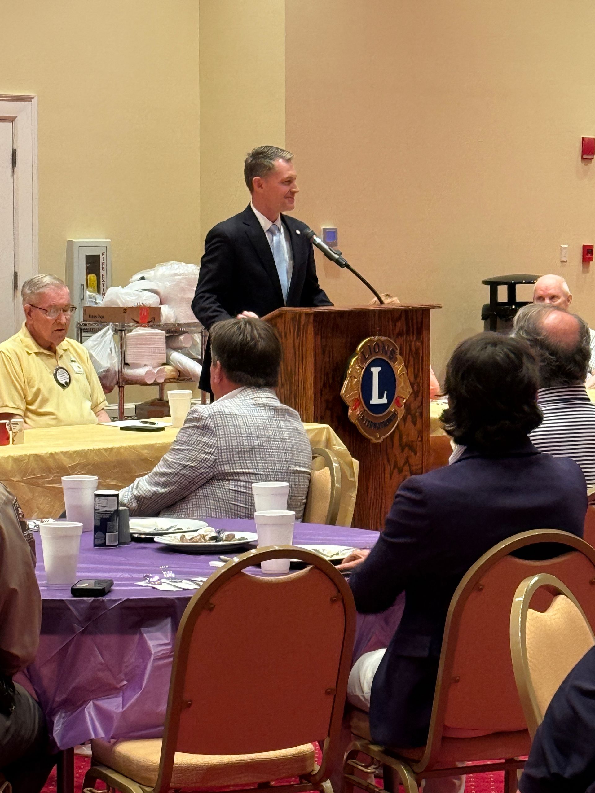 Man in suit speaks from a podium with Lions Club logo. People sit at tables in a room.