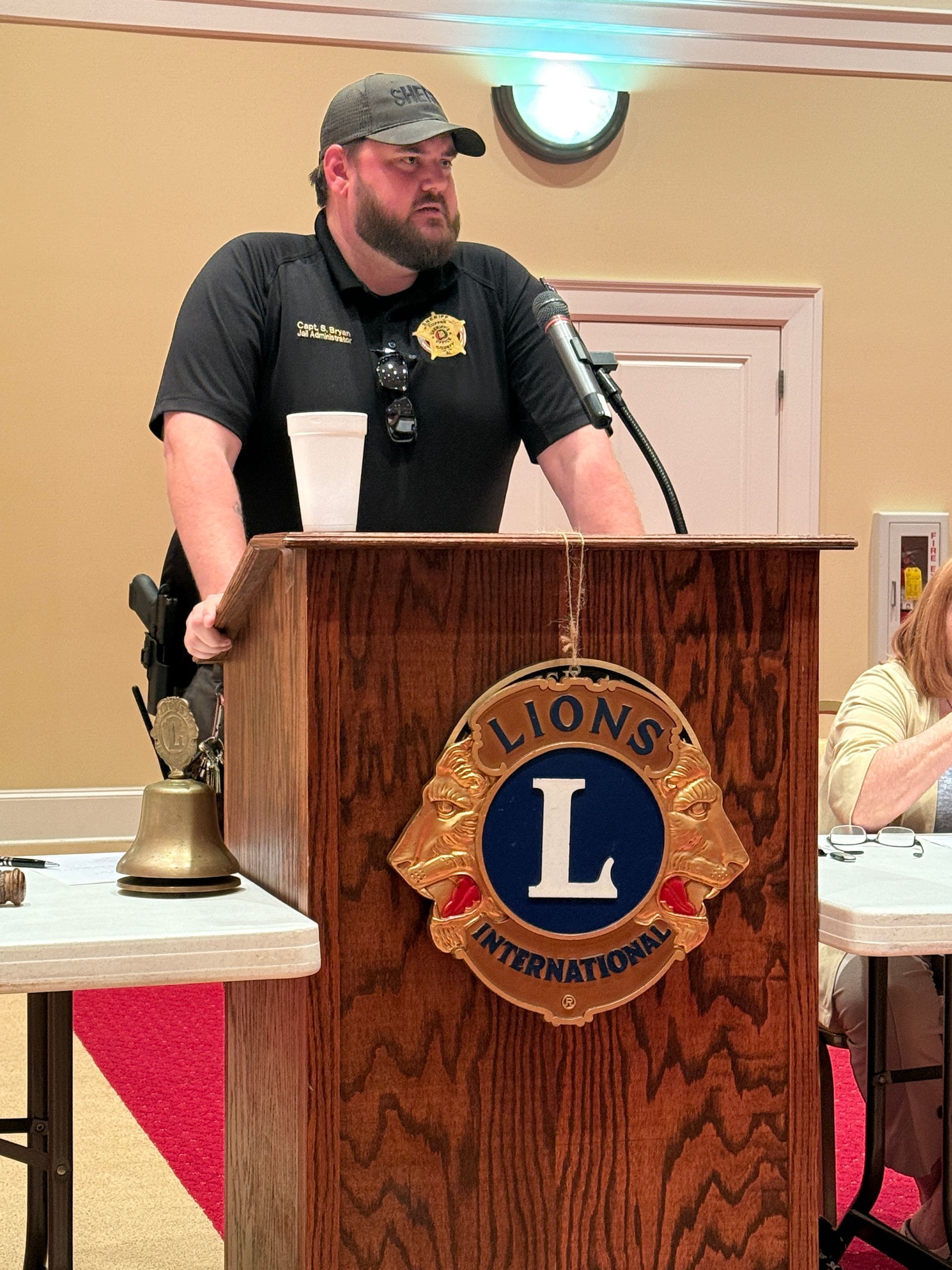 Man speaking at a Lions Club podium, wearing a dark uniform and hat.