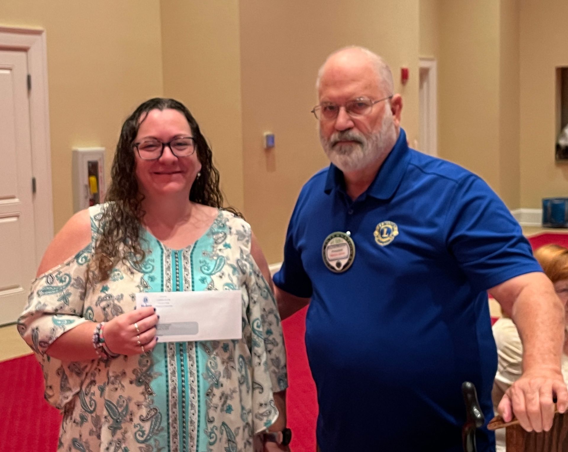 Woman in patterned top holds envelope, receiving check from man with blue shirt and Lions Club badge.