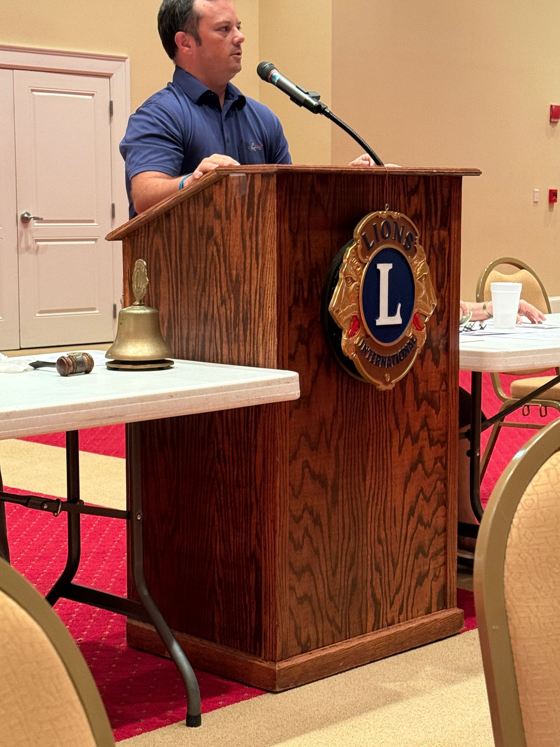 Man speaking at a Lions Club podium; wooden lectern with logo, bell, and table.