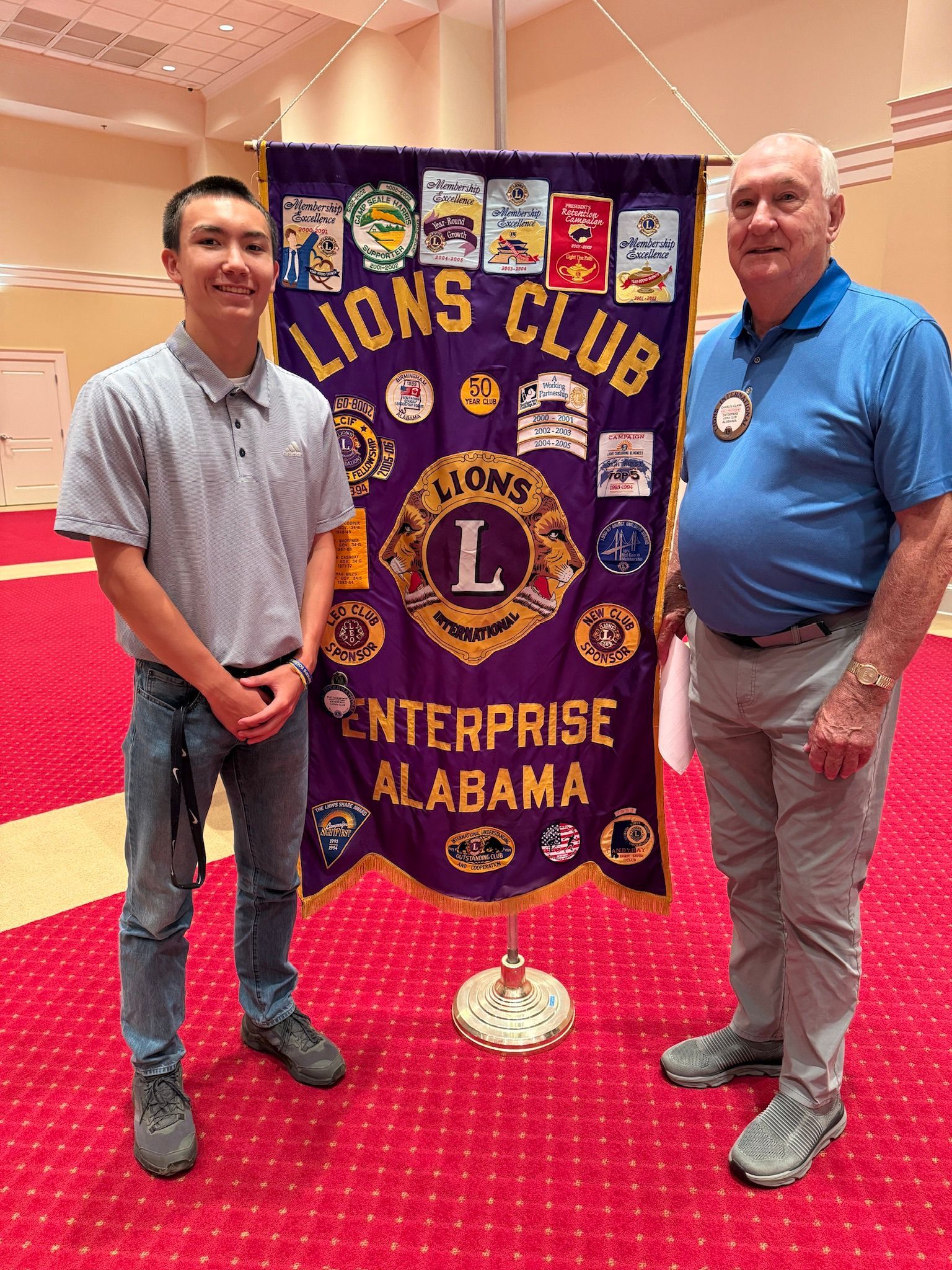 Two men standing in front of a Lions Club banner. The younger man wears jeans and a gray shirt; the older man has a blue polo.