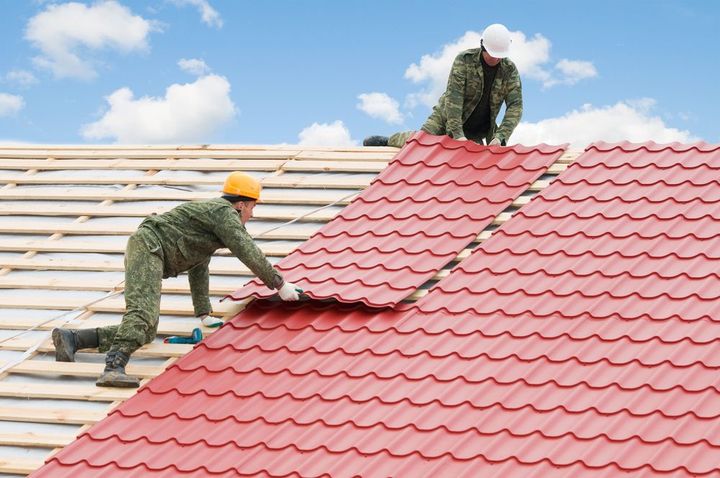 Two roofers installing red roof shingles on a house under a blue sky