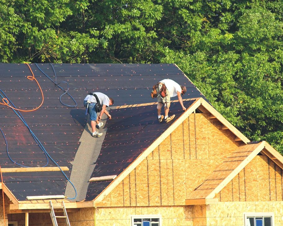 Two workers installing roofing on a house, with black underlayment and unfinished wood walls.