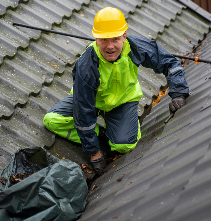 Worker in yellow hard hat and rain gear crouches on a wet corrugated roof during maintenance.