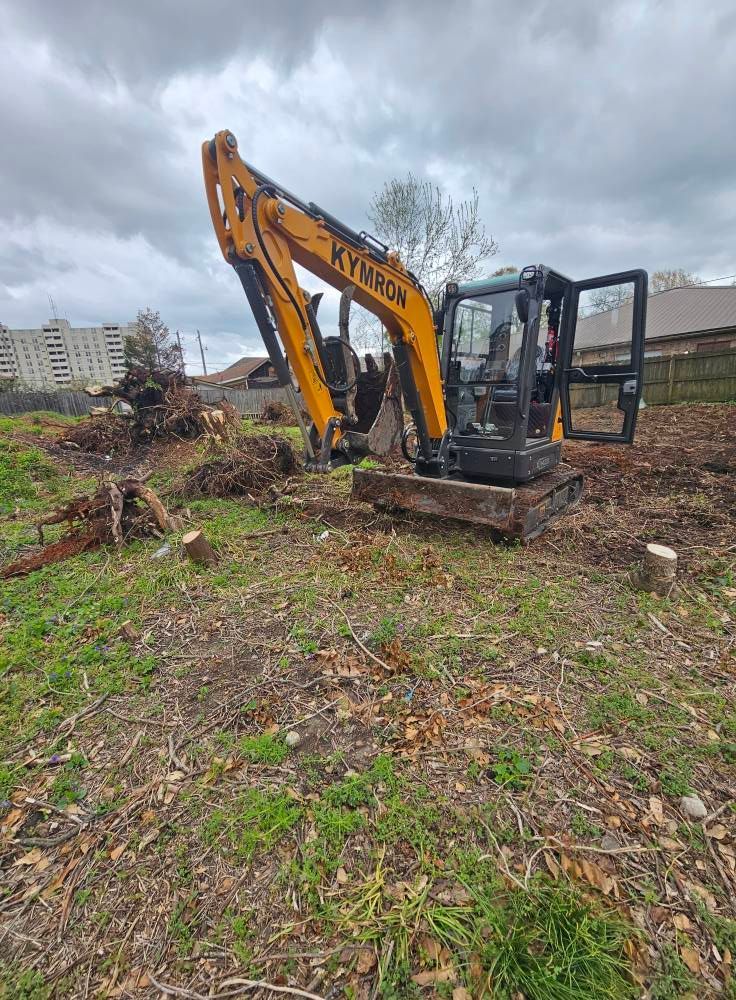 Yellow excavator clearing brush in a muddy field under a cloudy sky.