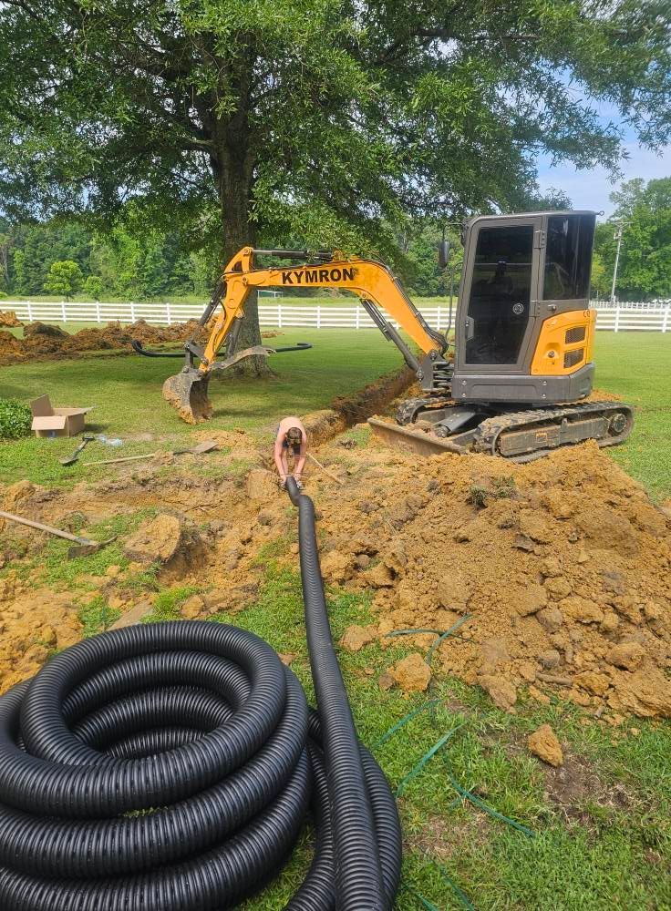 Excavator trenching a grassy yard with black drainage pipe coils in the foreground.