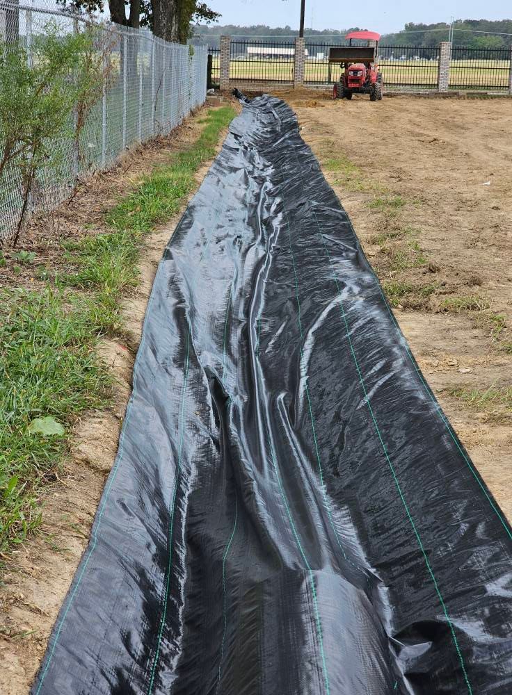 Black plastic liner in a long trench beside a fence, with a red tractor in the background.